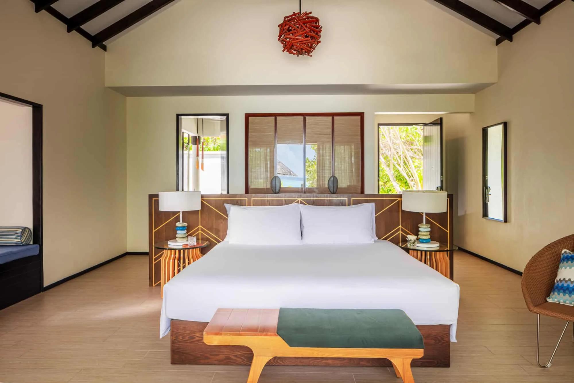 A Sunrise Beach Villa's bed beneath a vaulted timber-beamed ceiling and a hanging red driftwood pendant, with a sea view framed in a woven-blind window behind