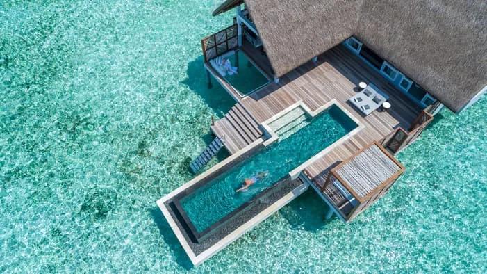 Aerial view directly over an overwater villa's plunge pool, a swimmer crossing the clear lagoon water below