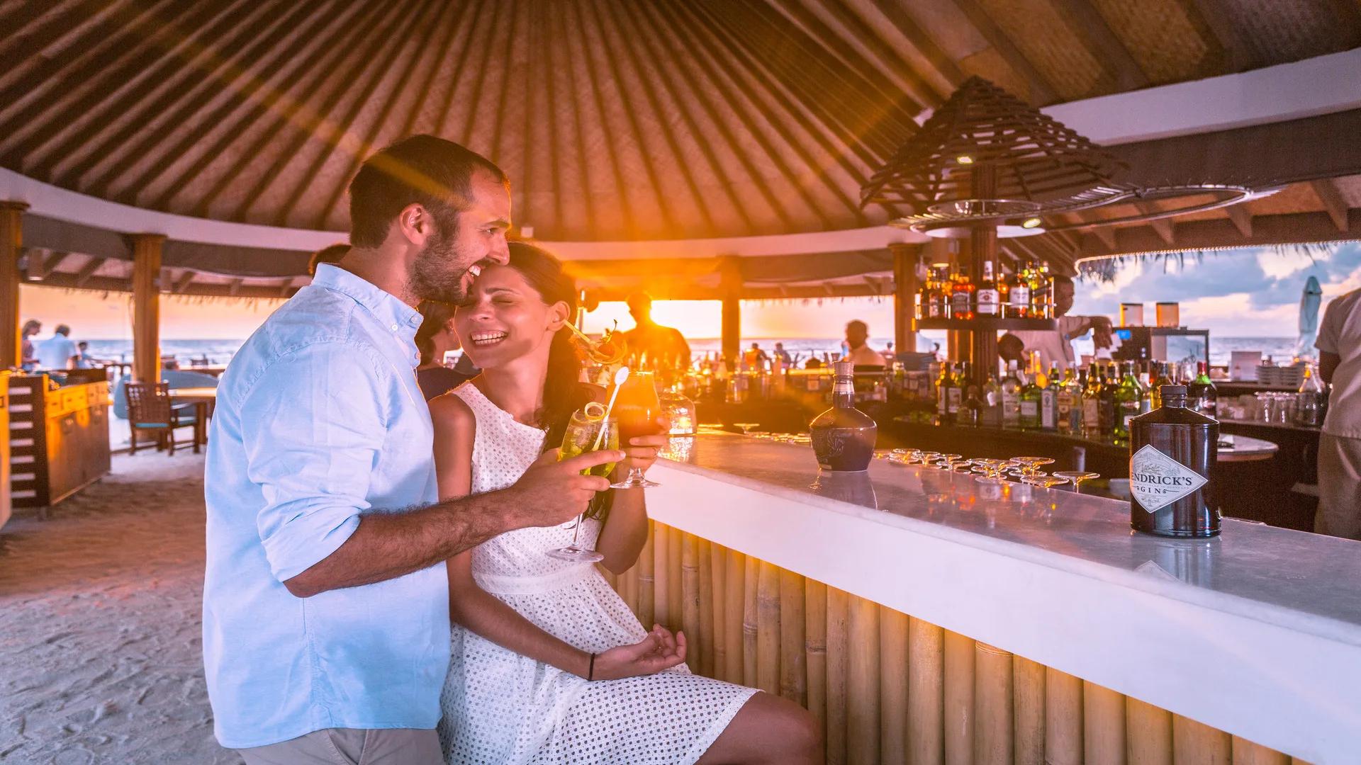 A couple sharing cocktails at Huvan's circular thatched bar at sunset, the bartender pouring drinks and the beach visible beyond