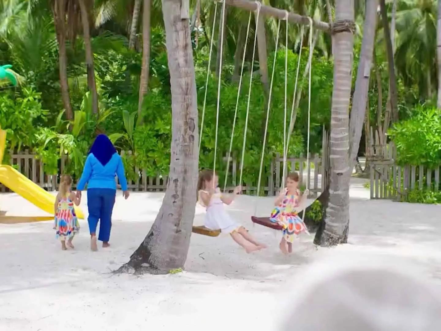 Children on rope swings under palm trees at Amilla's Kids Club, a staff member walking with another child nearby
