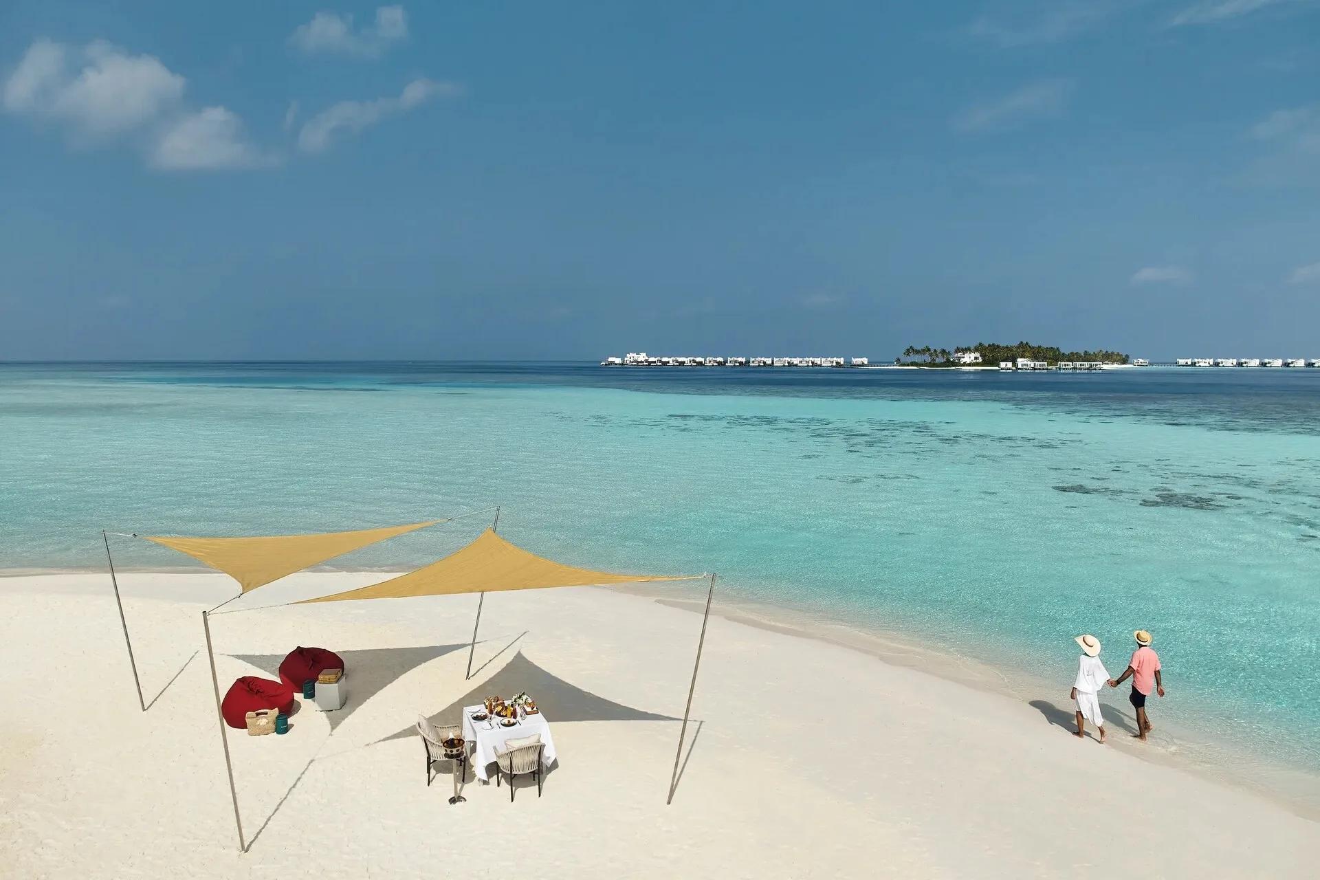 Aerial view of a private dining setup with a shade canopy on a stretch of sand, a couple walking along the shoreline nearby and the resort's overwater villas visible across the lagoon