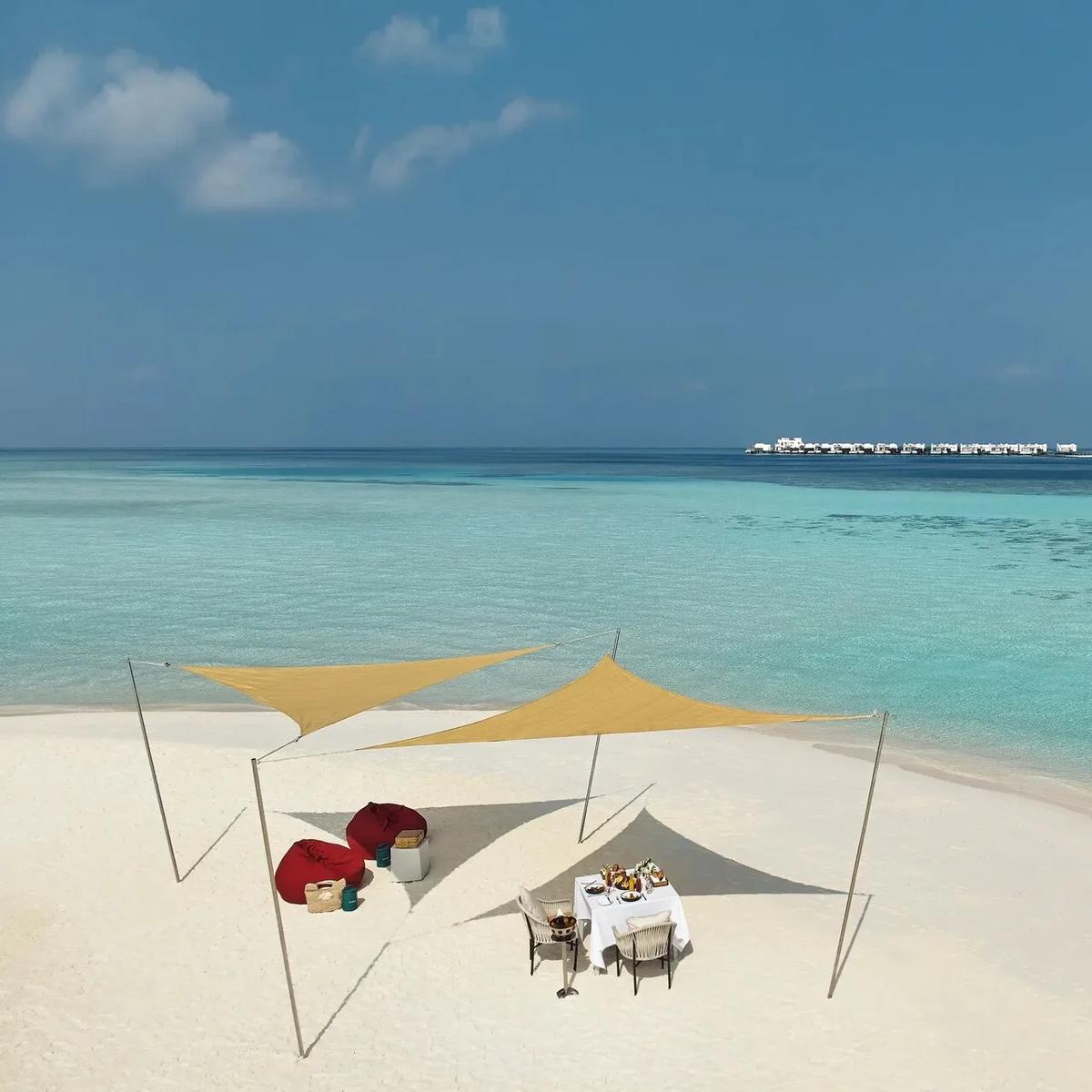 Aerial view of a private dining setup with a shade canopy on a stretch of sand, a couple walking along the shoreline nearby and the resort's overwater villas visible across the lagoon