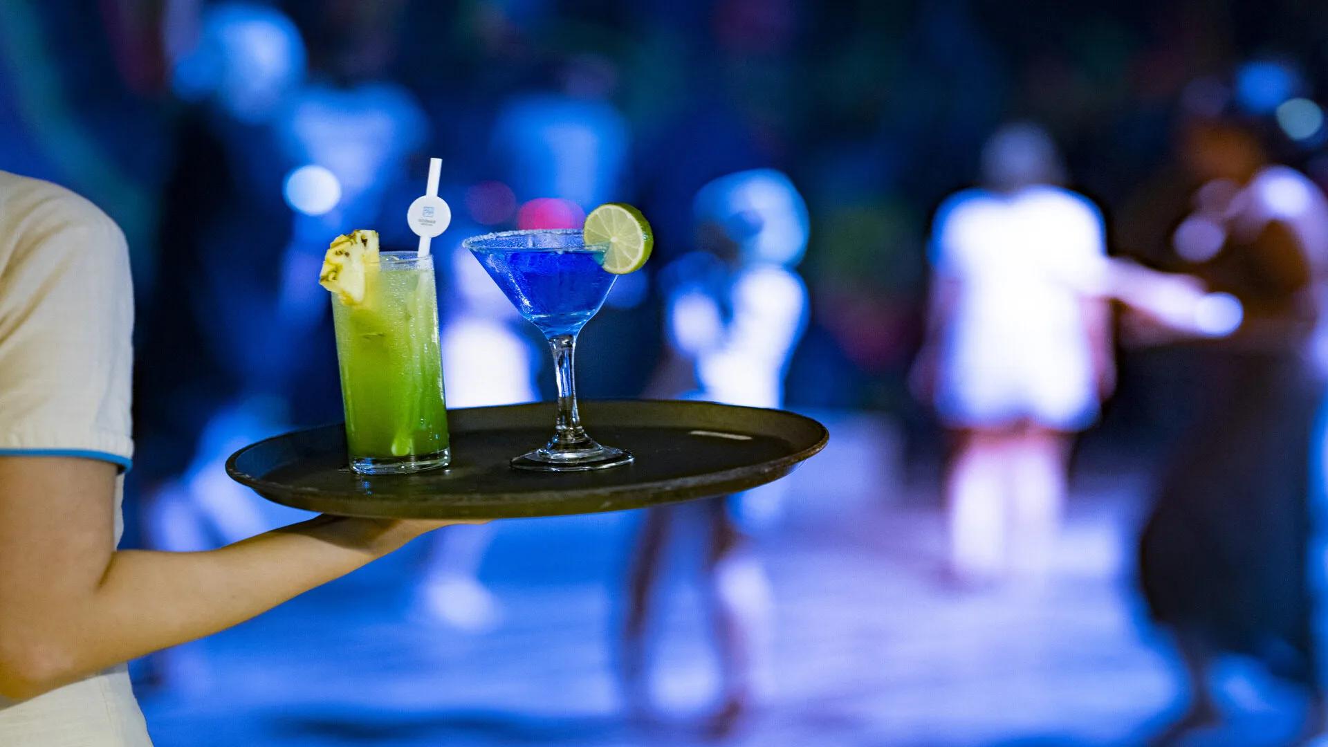Two cocktails on a tray carried across Sand Bar's dance floor, dancers blurred in the blue disco light behind