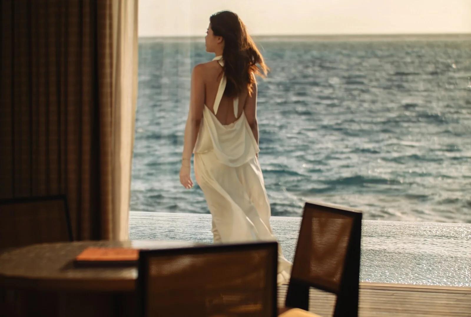 A guest in a white dress standing at the edge of an Ocean Villa's overwater terrace at sunset, open water ahead