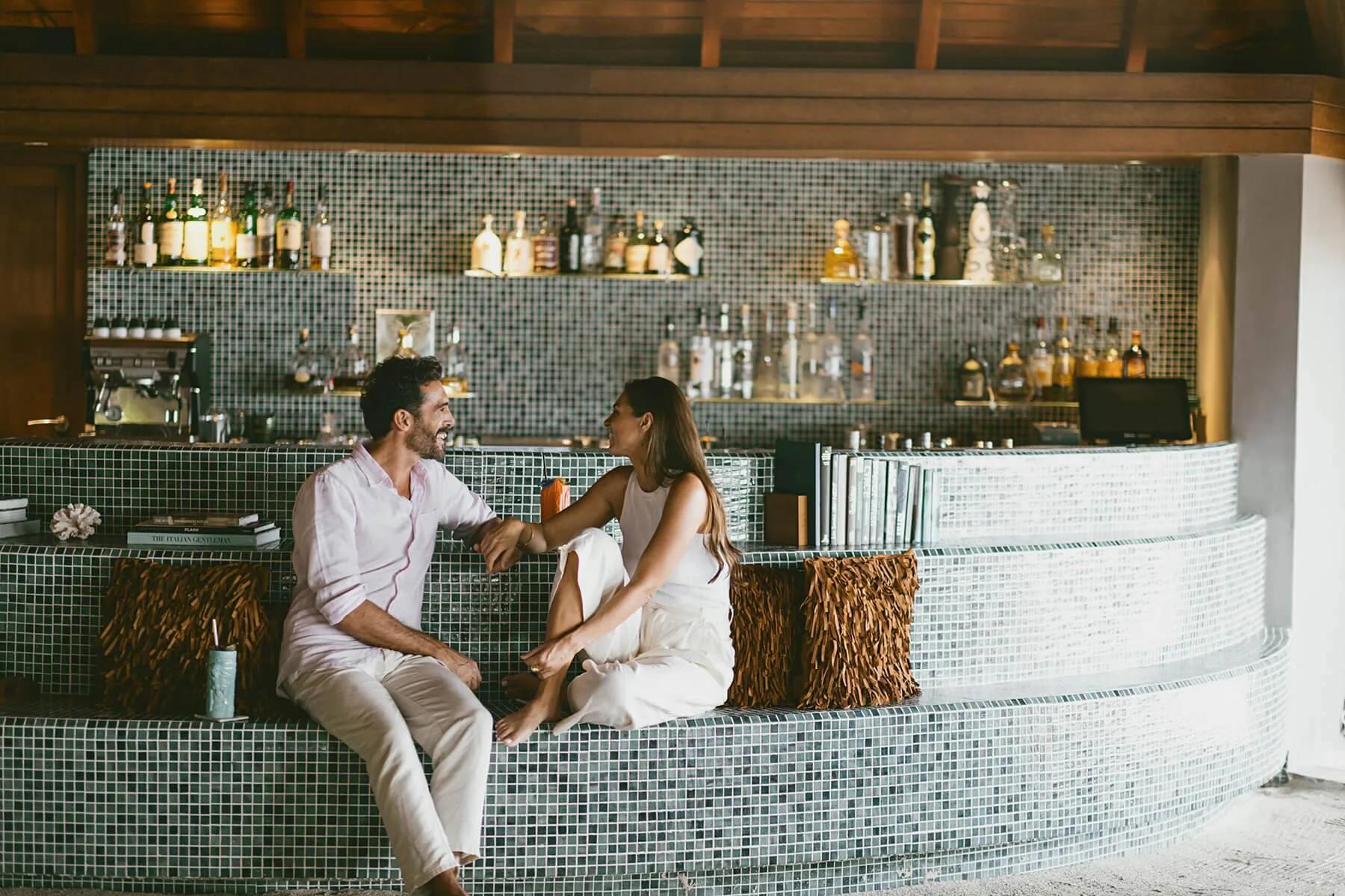 A couple seated at Umbar's mosaic-tiled bar counter, shelves of spirits lit behind them