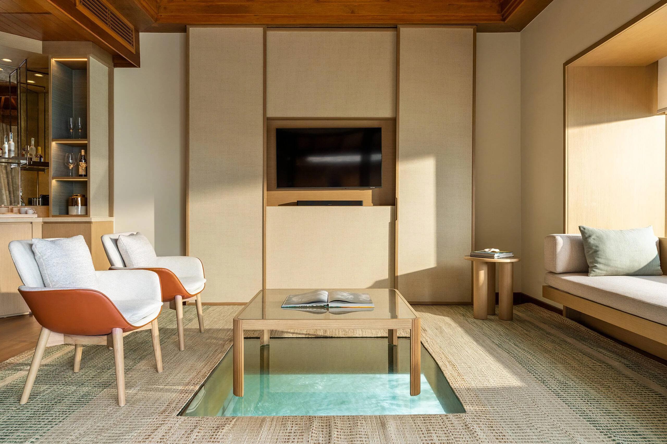 The living room of an Ocean Bungalow with Pool, a glass-topped table set over a sunken water feature, with a bar nook and lounge chairs behind