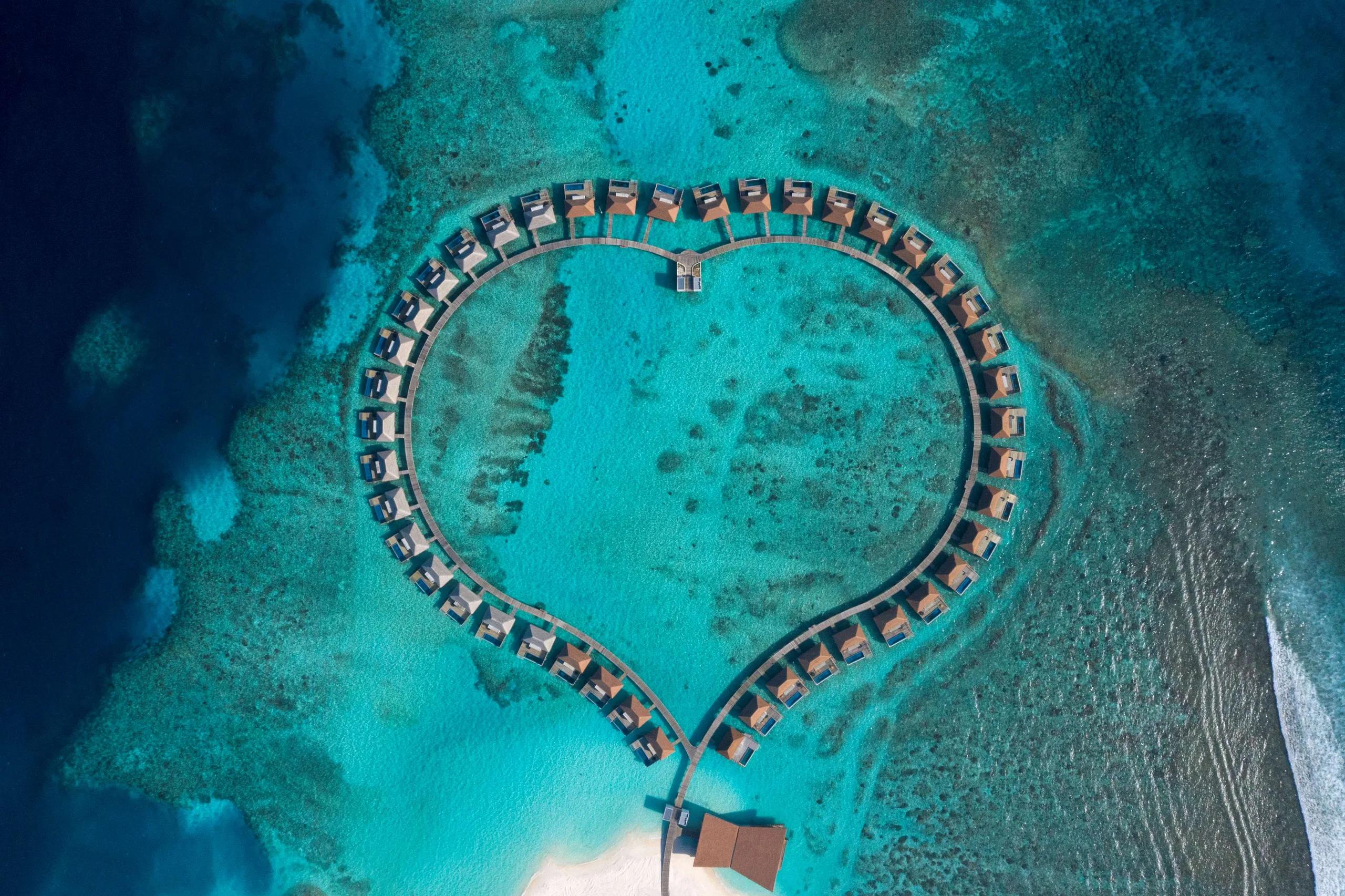 A heart-shaped loop of overwater villas at Radisson Blu Resort Maldives, seen directly from above, encircling a private stretch of lagoon within the reef