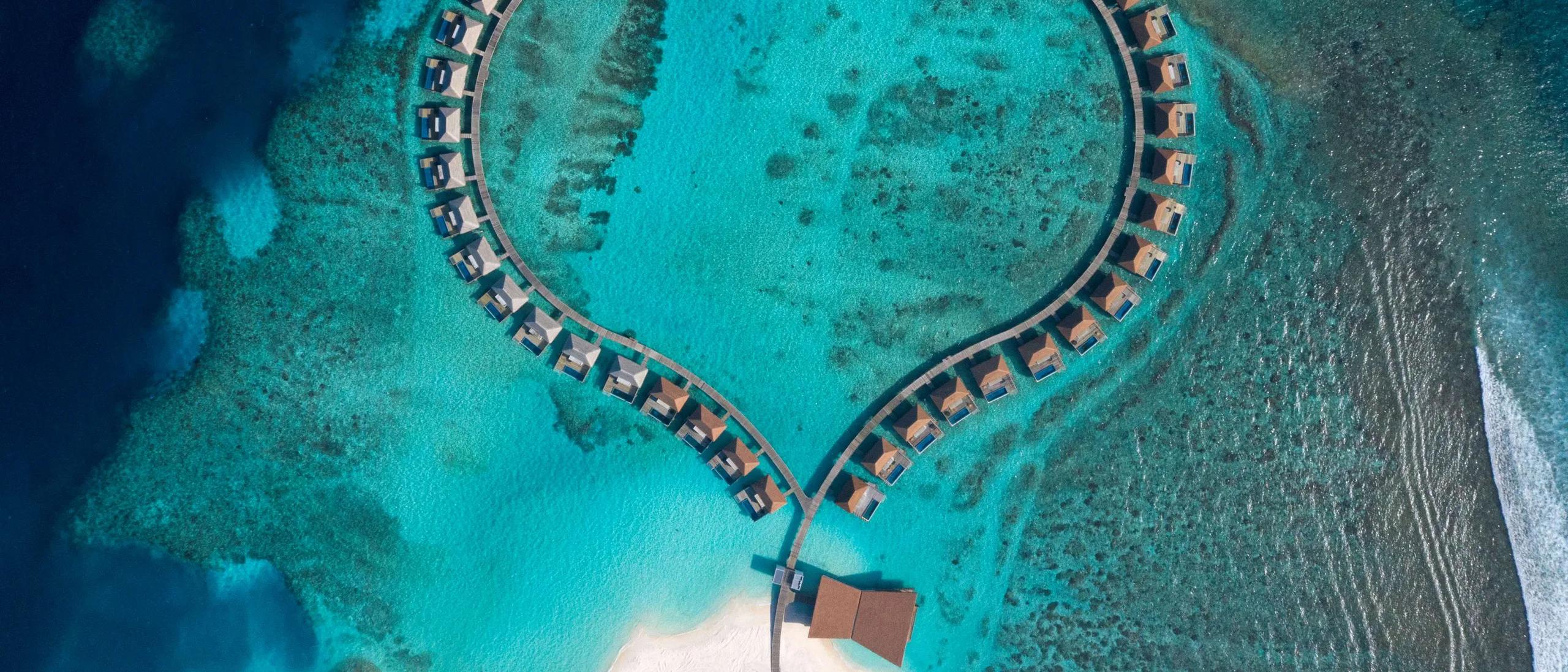 A heart-shaped loop of overwater villas at Radisson Blu Resort Maldives, seen directly from above, encircling a private stretch of lagoon within the reef