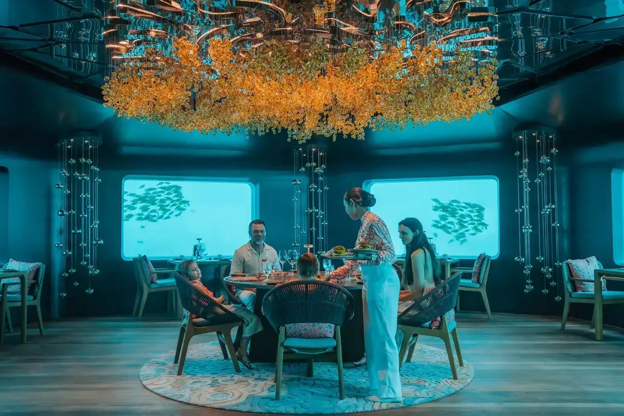 A family dining at Bubble, seated beneath a gold chandelier as reef fish pass the panoramic windows