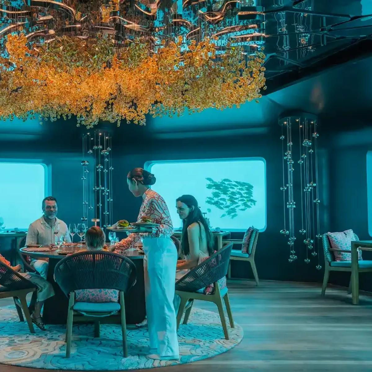 A family dining at Bubble, seated beneath a gold chandelier as reef fish pass the panoramic windows