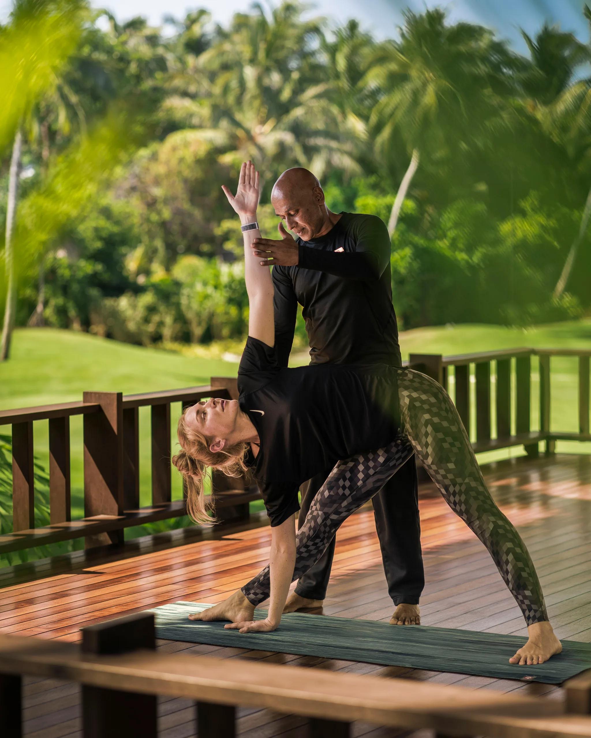 An instructor guides a guest through a yoga pose on an outdoor deck, palm trees and a golf fairway behind them