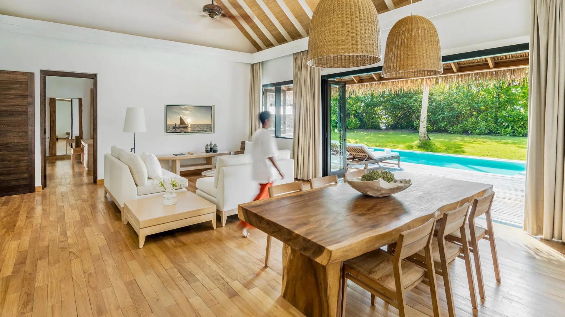 Maalifushi Beach Villa's living and dining room opening onto its private pool and lawn, staff member walking through