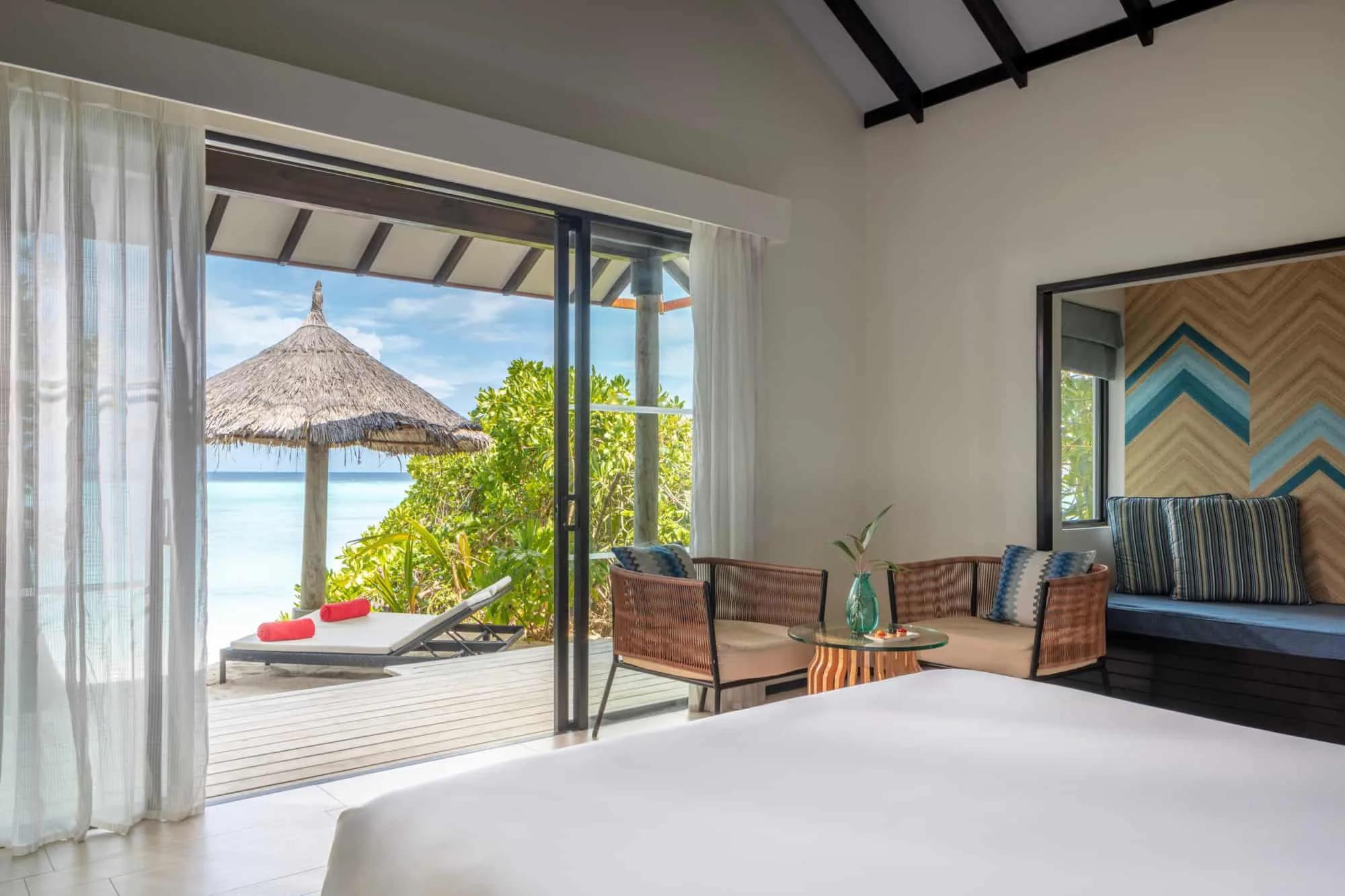 A thatched-roof beach cabana framing a view of the lagoon through palm fronds, with rattan chairs and sun loungers set out on the sand