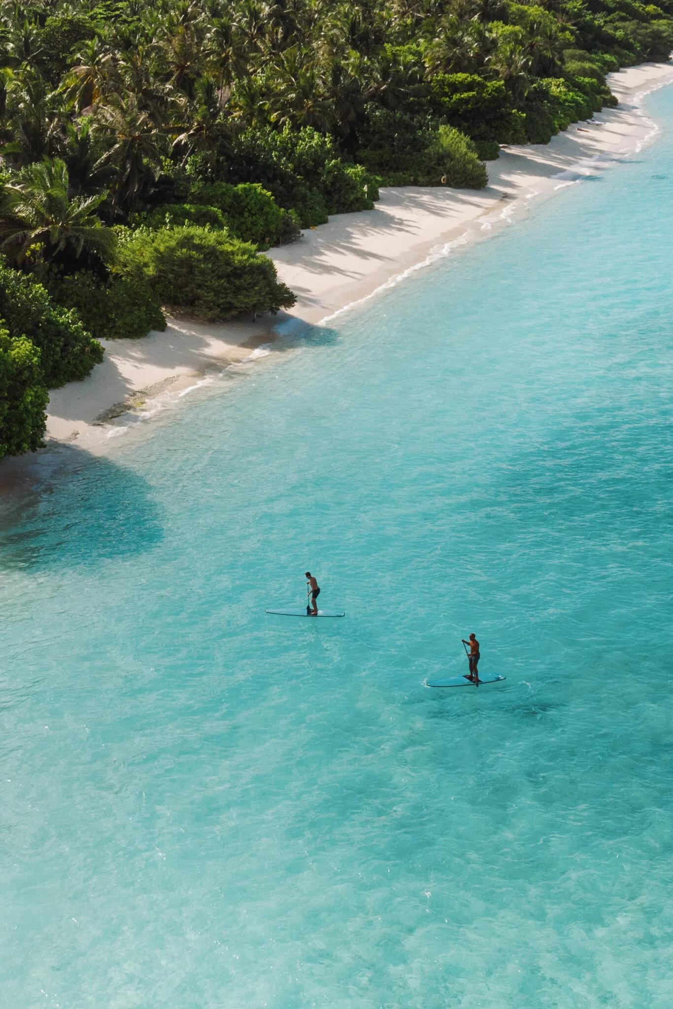 Two guests paddleboard along a shallow turquoise lagoon, a white-sand beach and dense jungle running the length of the shore beside them