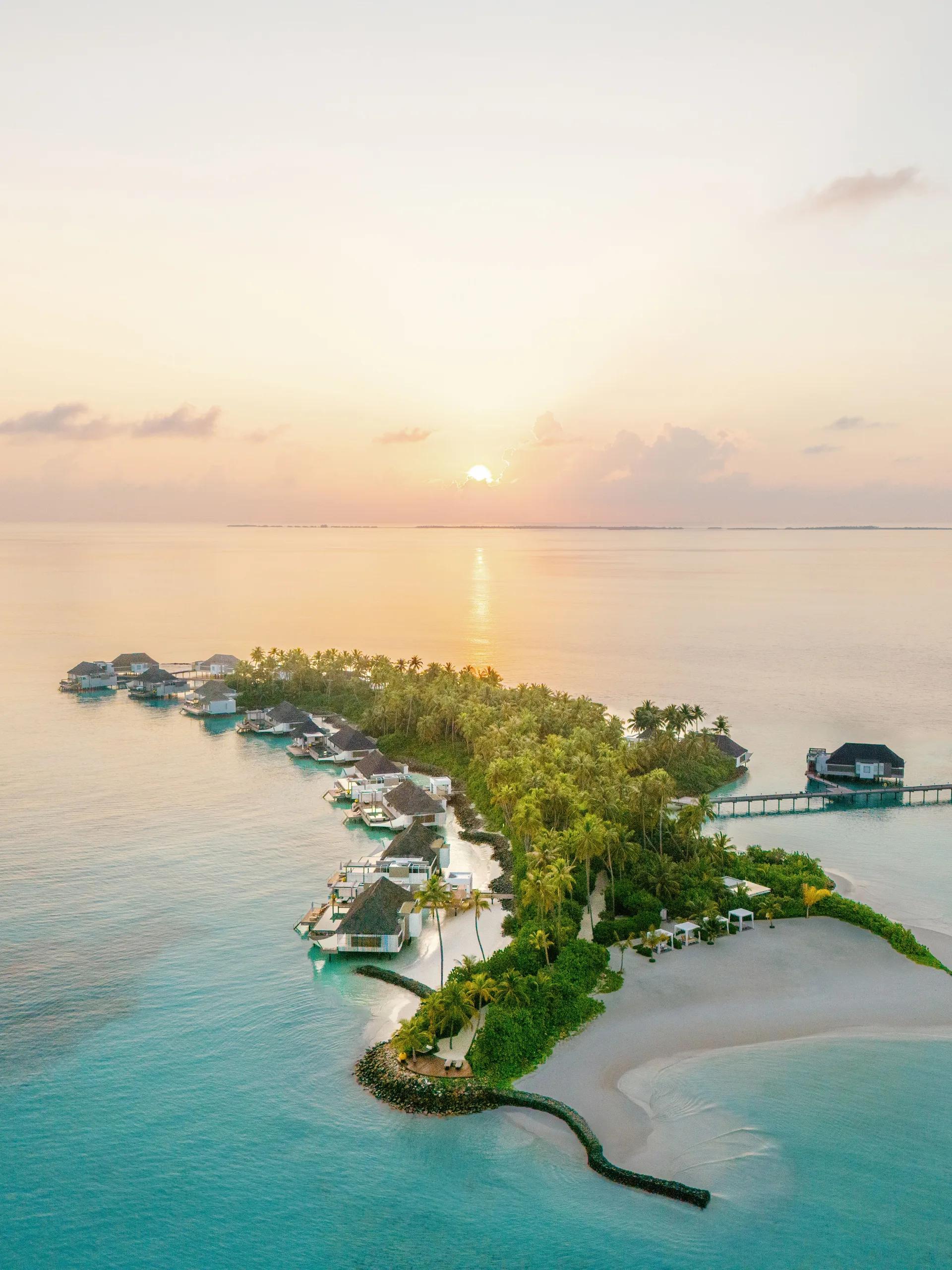 Aerial view of Cheval Blanc Randheli at dusk, overwater villas tracing the shoreline into Noonu Atoll's lagoon