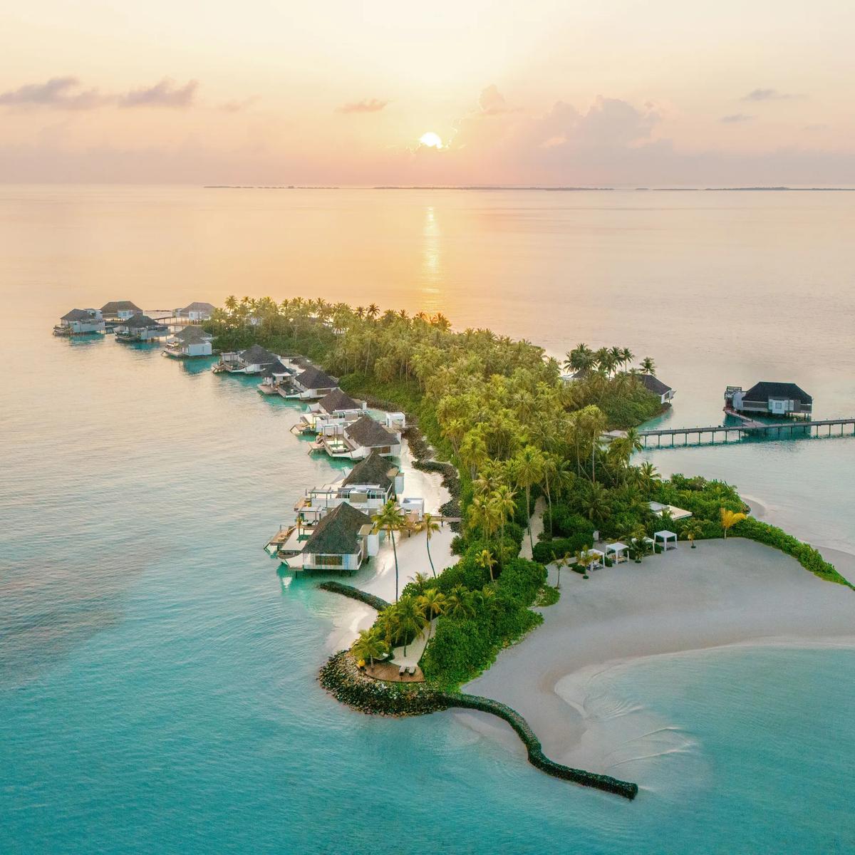 Aerial view of Cheval Blanc Randheli at dusk, overwater villas tracing the shoreline into Noonu Atoll's lagoon