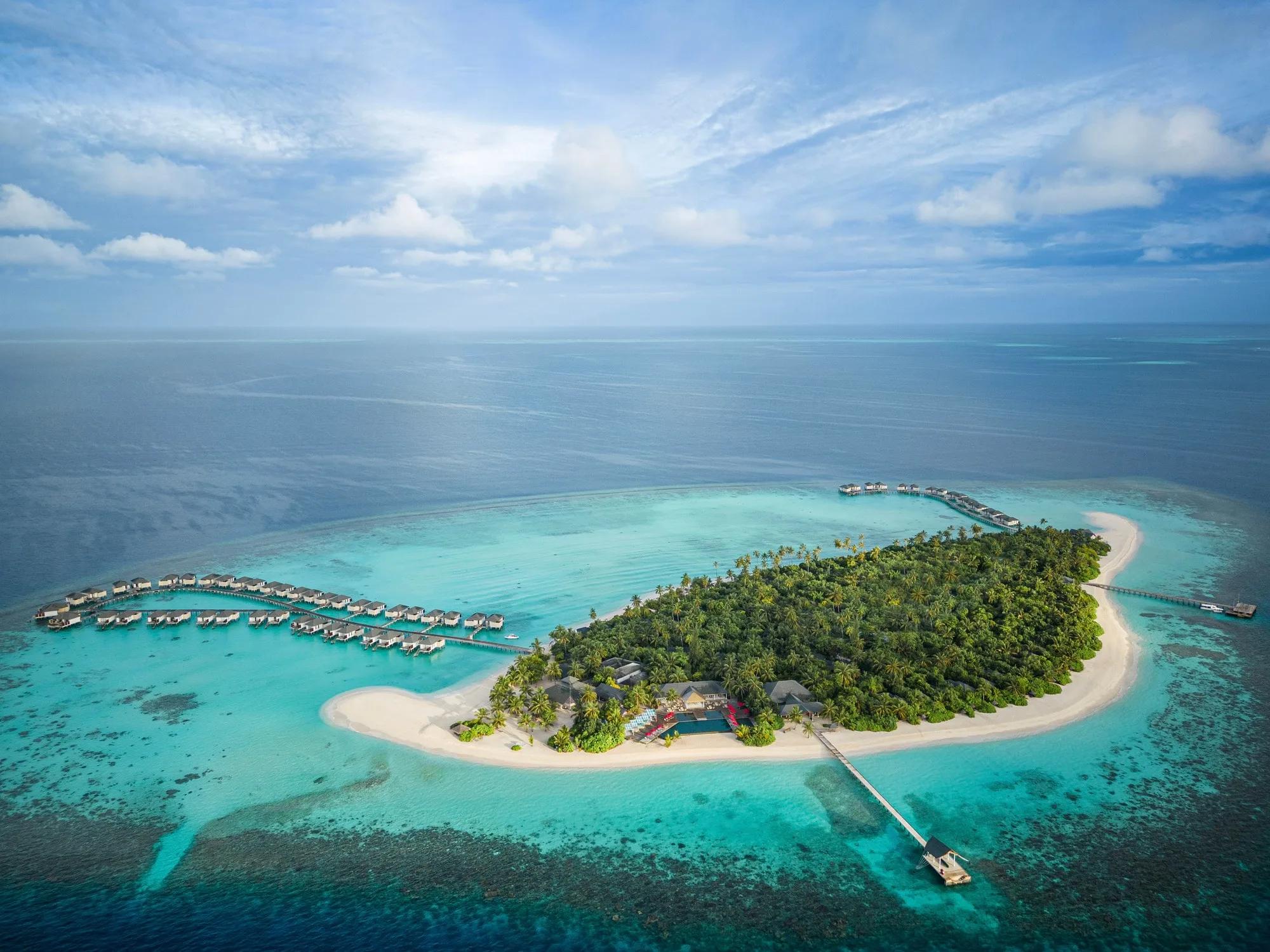 Wide aerial view of Havodda's teardrop-shaped island, ringed by reef, with two rows of overwater villas on jetties extending from either side