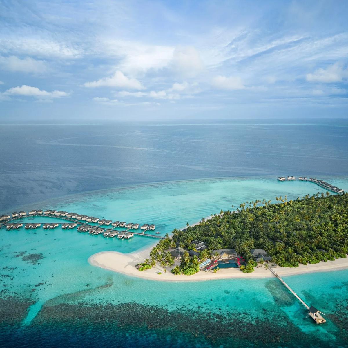 Wide aerial view of Havodda's teardrop-shaped island, ringed by reef, with two rows of overwater villas on jetties extending from either side