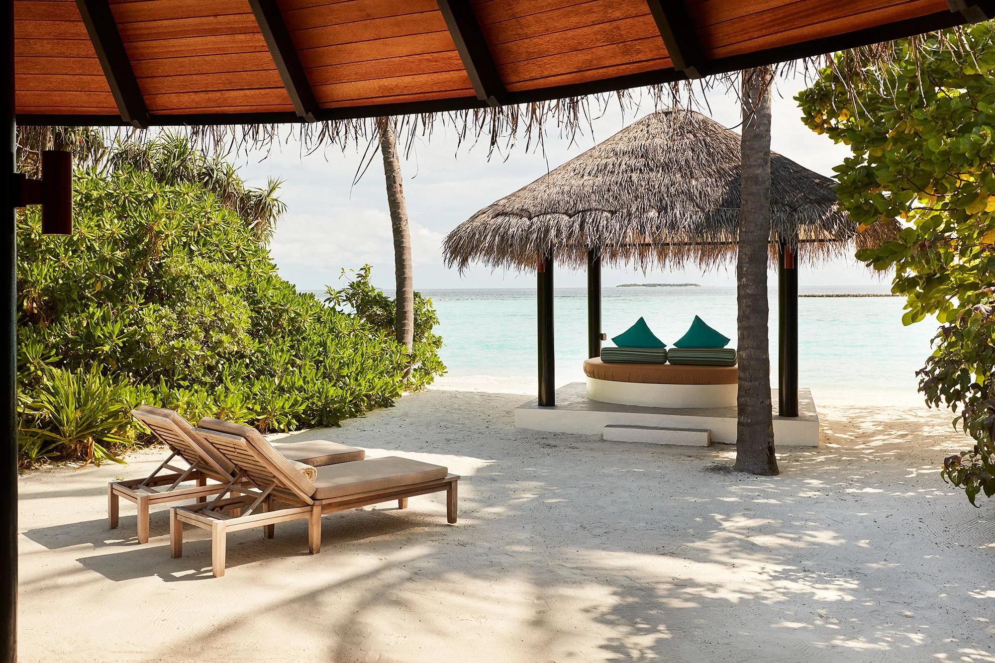A thatched beach sala with teal cushions framed by palm fronds, open to the lagoon at a Deluxe Beach Villa with Pool