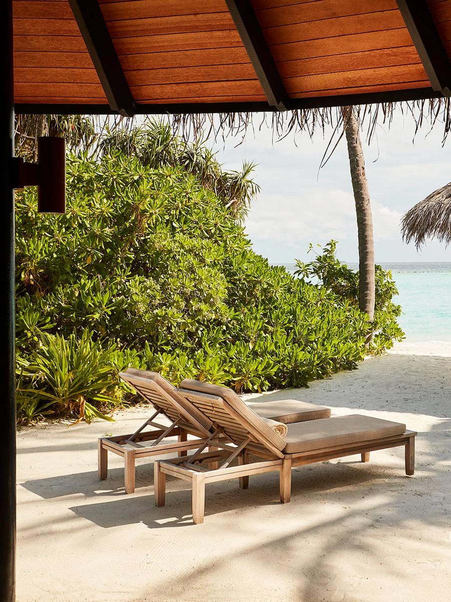 A thatched beach sala with teal cushions framed by palm fronds, open to the lagoon at a Deluxe Beach Villa with Pool