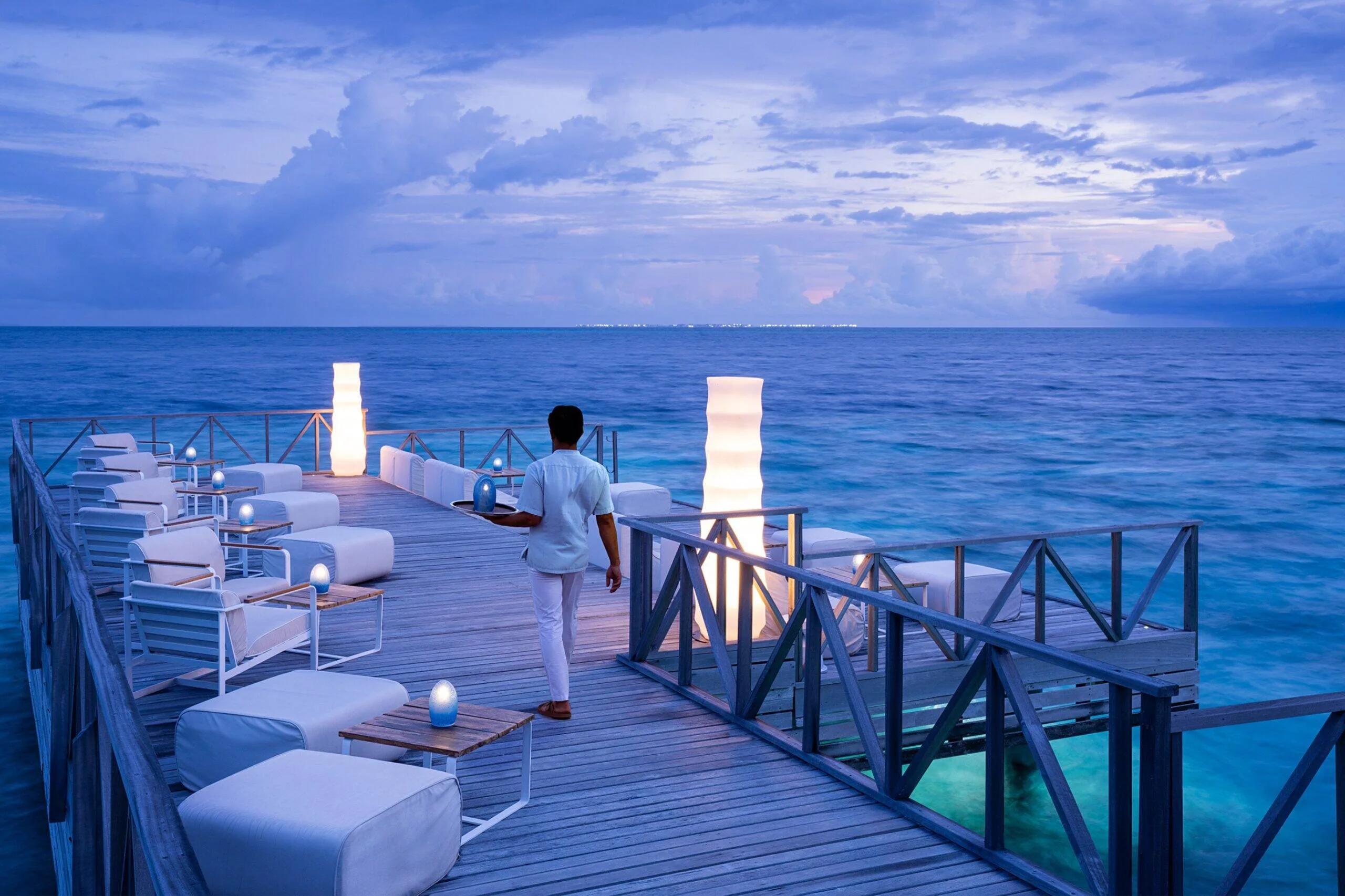 A waiter carrying a drinks tray along Raw Sunset Deck's lantern-lit deck at dusk