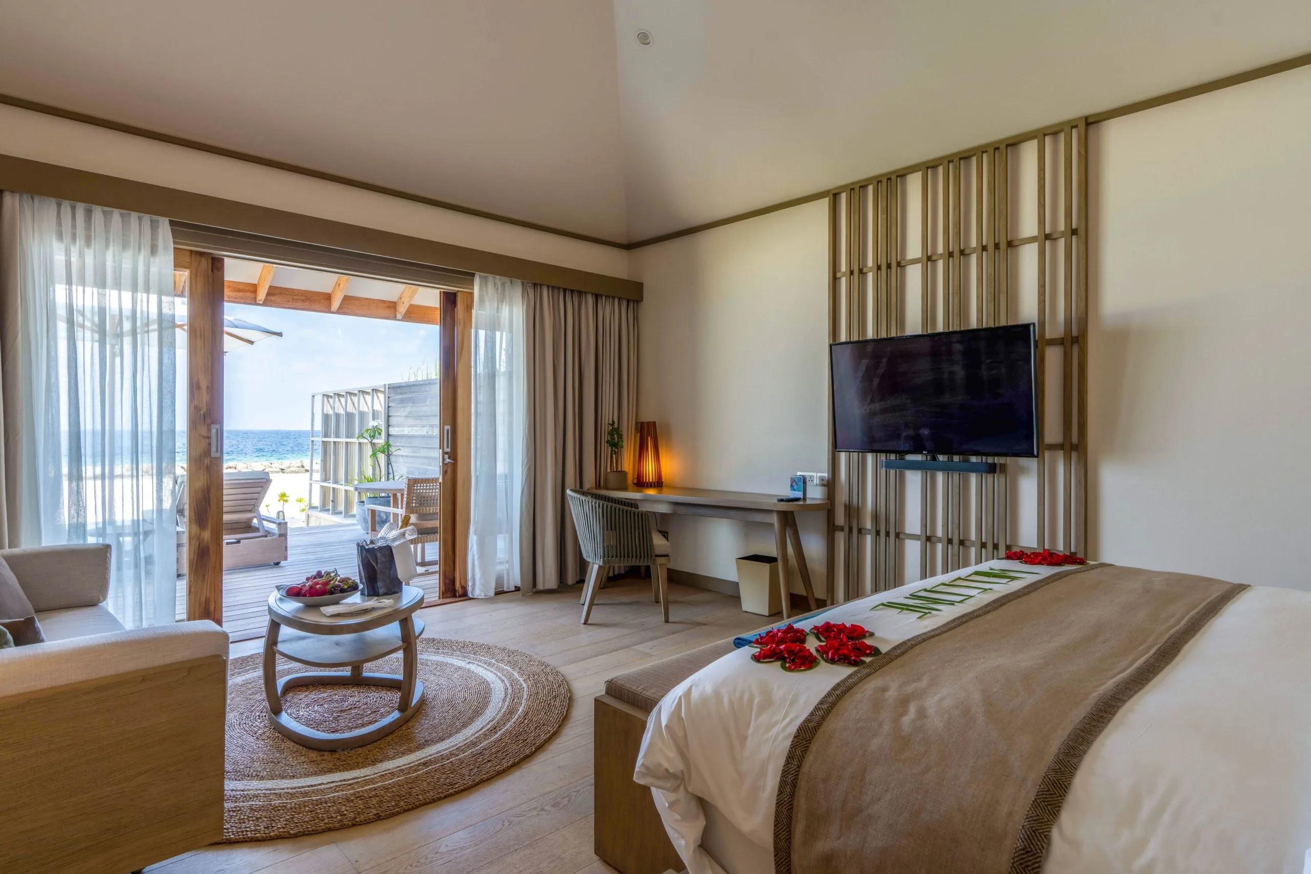 A beach villa bedroom with rose petals arranged on the bed, sliding doors open to a deck with sun loungers and the ocean beyond