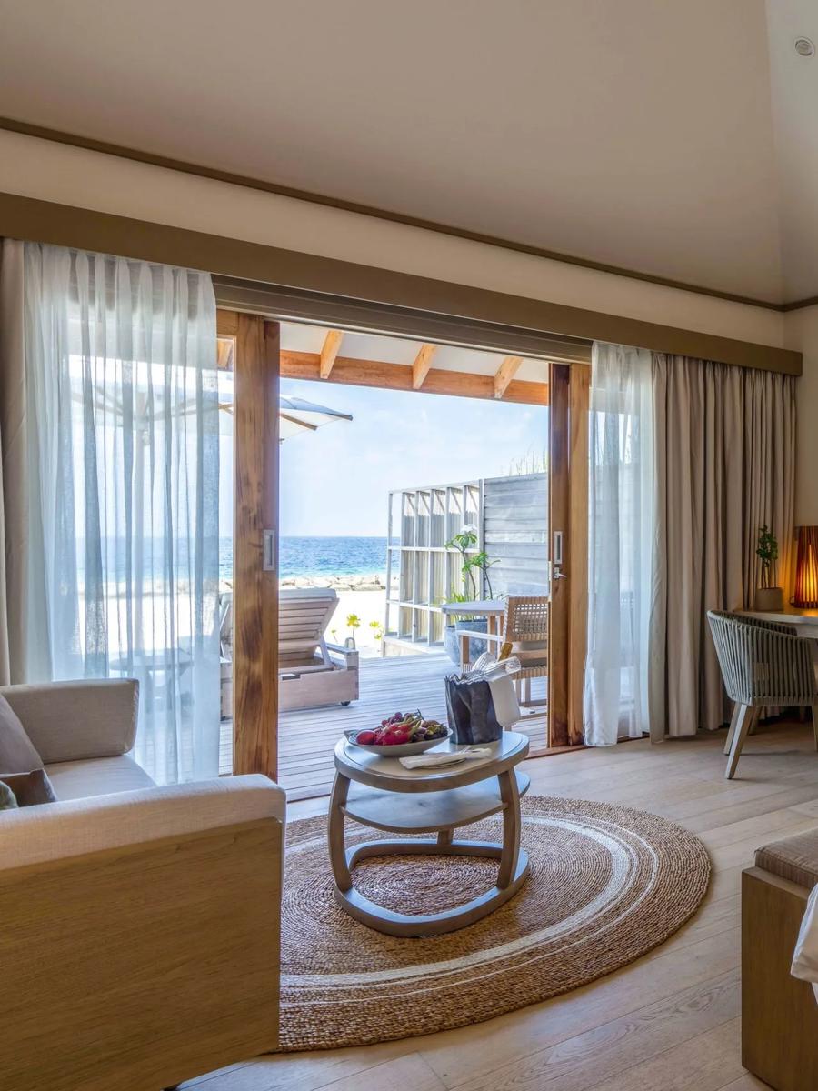 A beach villa bedroom with rose petals arranged on the bed, sliding doors open to a deck with sun loungers and the ocean beyond