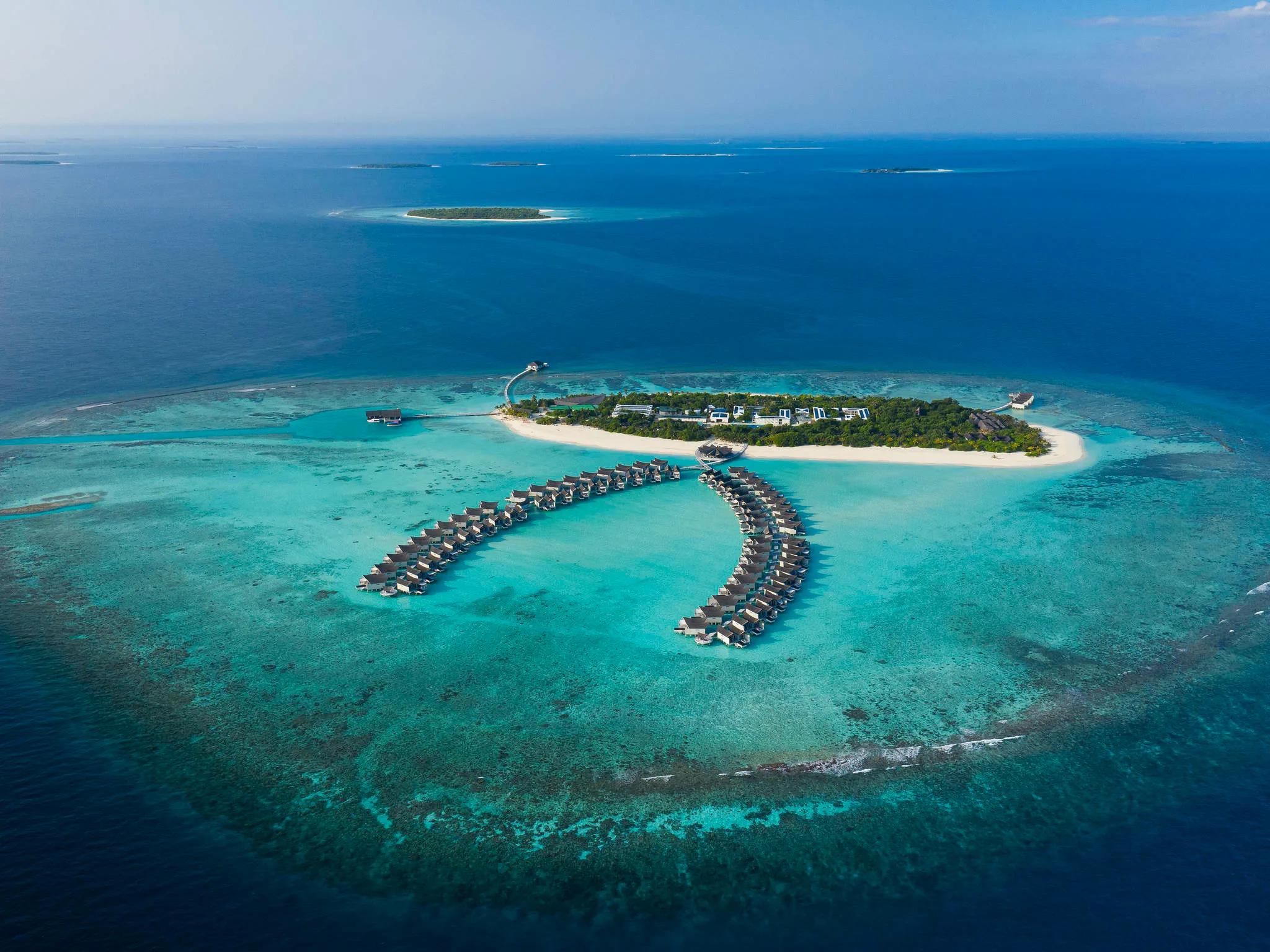 Aerial view of Kuredhivaru Resort & Spa Maldives, its curved double row of overwater villas extending from the beach into the lagoon, reef and open ocean beyond