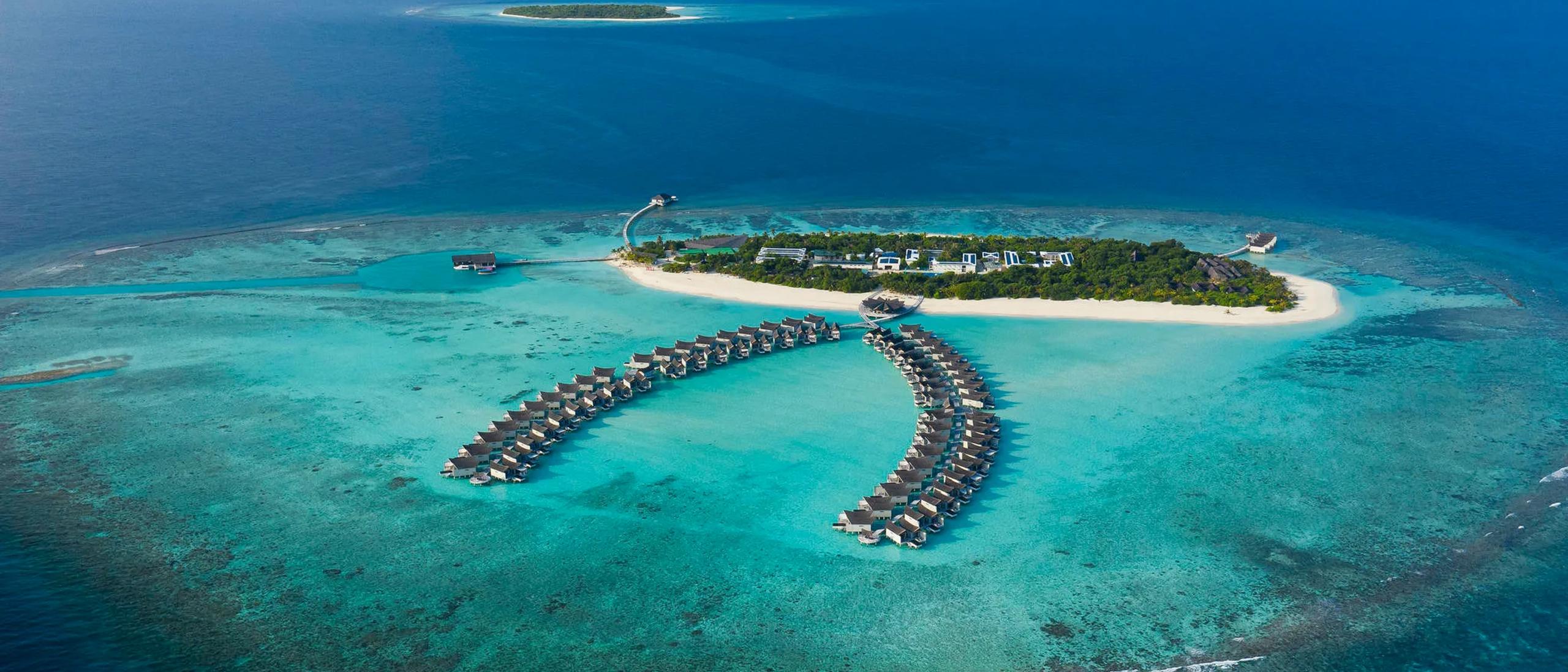 Aerial view of Kuredhivaru's island and its two curved rows of overwater villas reaching into the lagoon, reef visible in the surrounding water