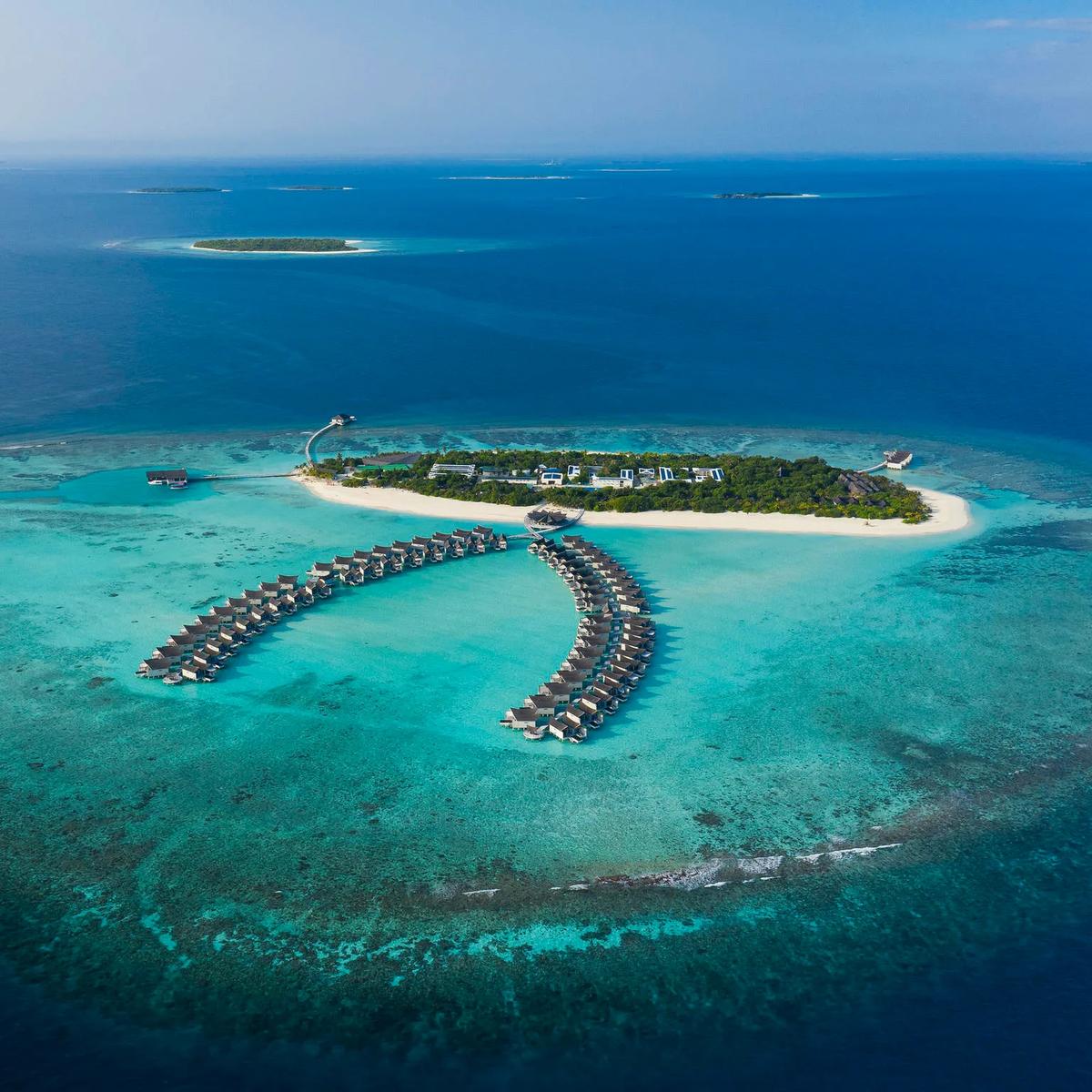 Aerial view of Kuredhivaru Resort & Spa Maldives, its curved double row of overwater villas extending from the beach into the lagoon, reef and open ocean beyond