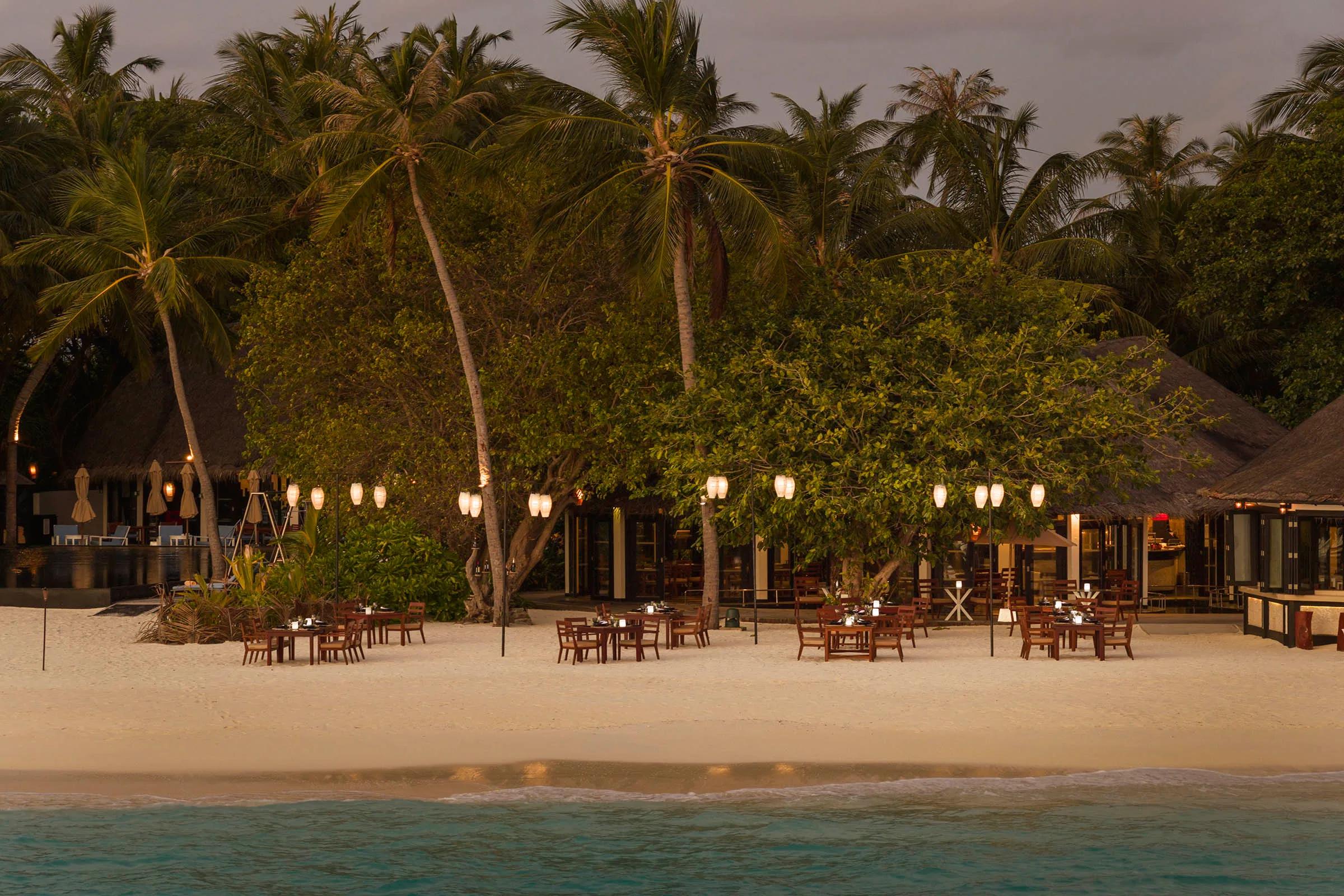 Lantern-lit dining tables set on the beach under palm trees at Islander's Grill in the evening