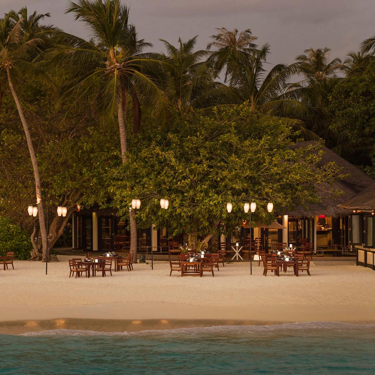 Lantern-lit dining tables set on the beach under palm trees at Islander's Grill in the evening