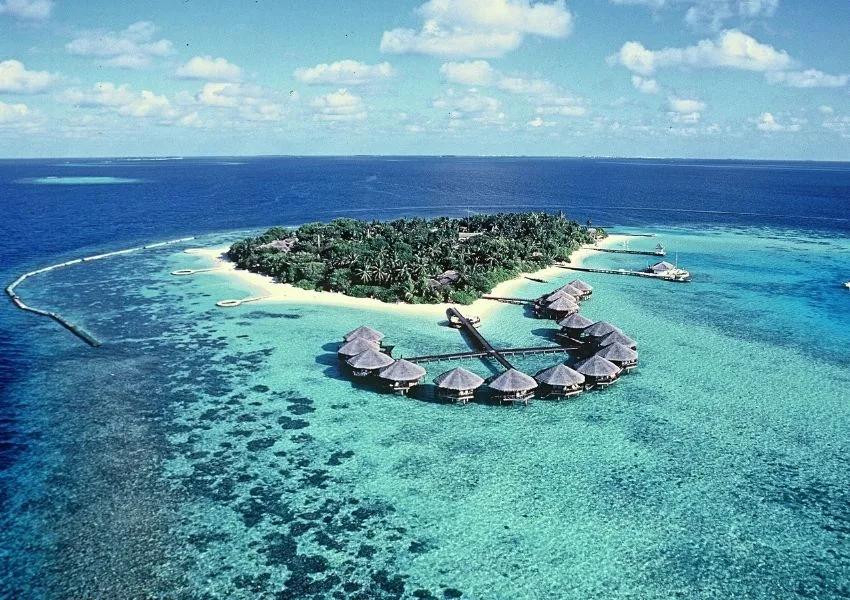 Aerial view of Baros Maldives — the reef-ringed island with overwater villas fanning out from its point and a sandy beach curving along the shore