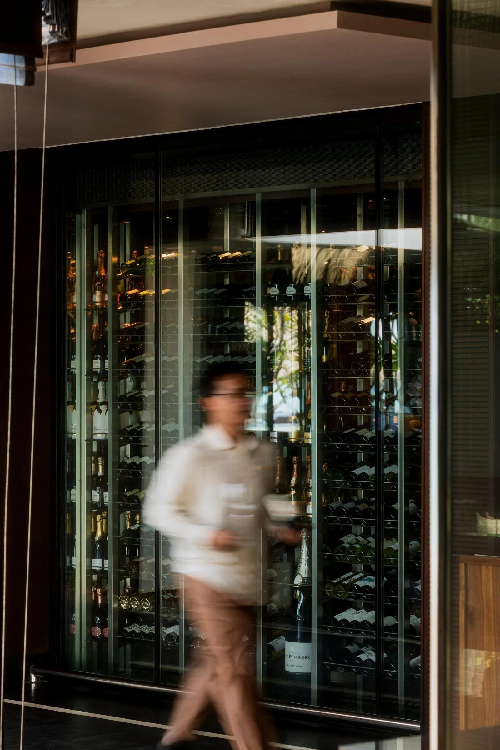 A staff member walks past Reethi's glass-walled wine cellar at One&Only Reethi Rah, carrying a bottle of champagne