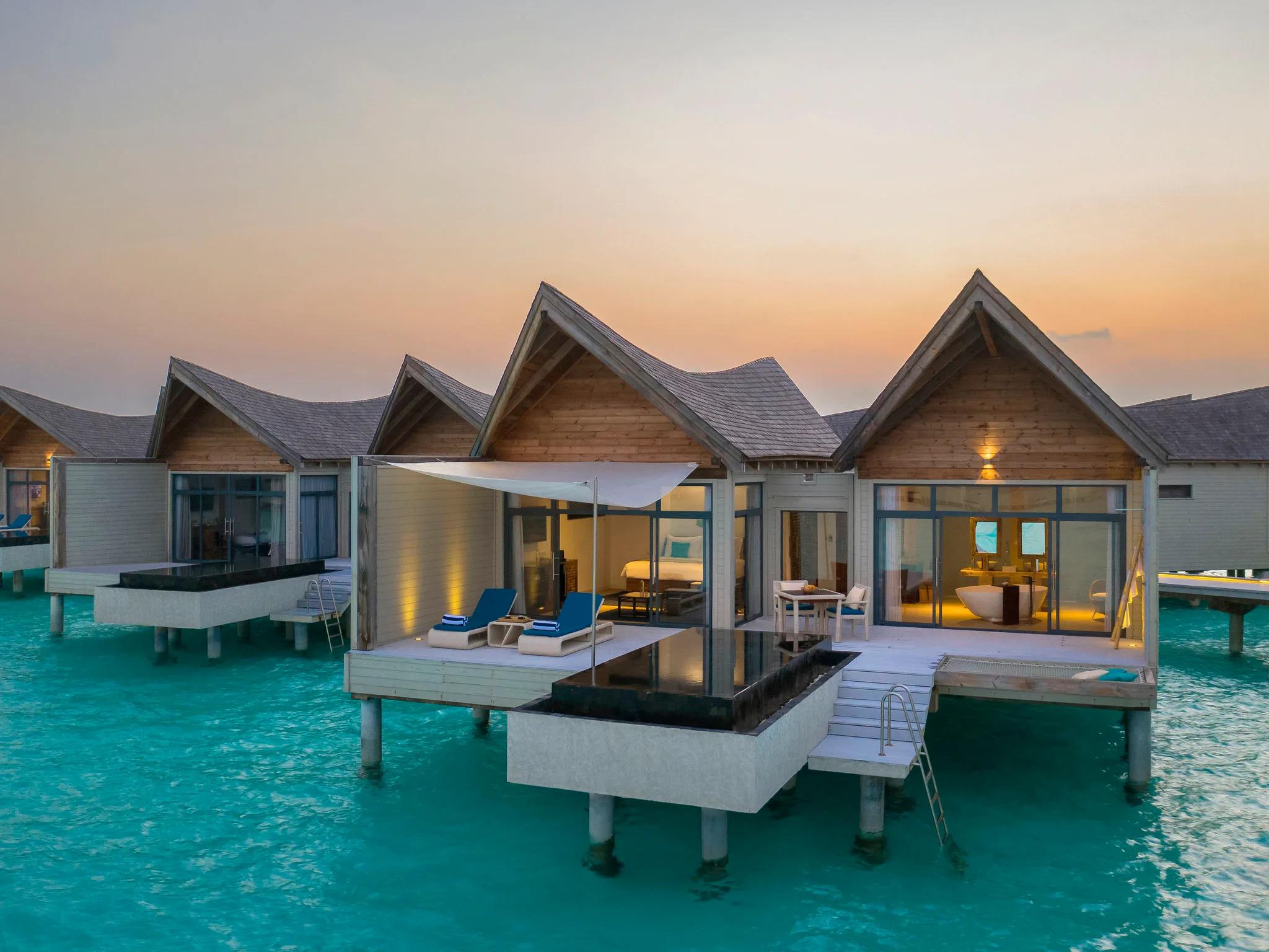Exterior view of an overwater pool villa at dusk, gabled wood roofs and a private plunge pool on the deck, steps leading down into the lagoon