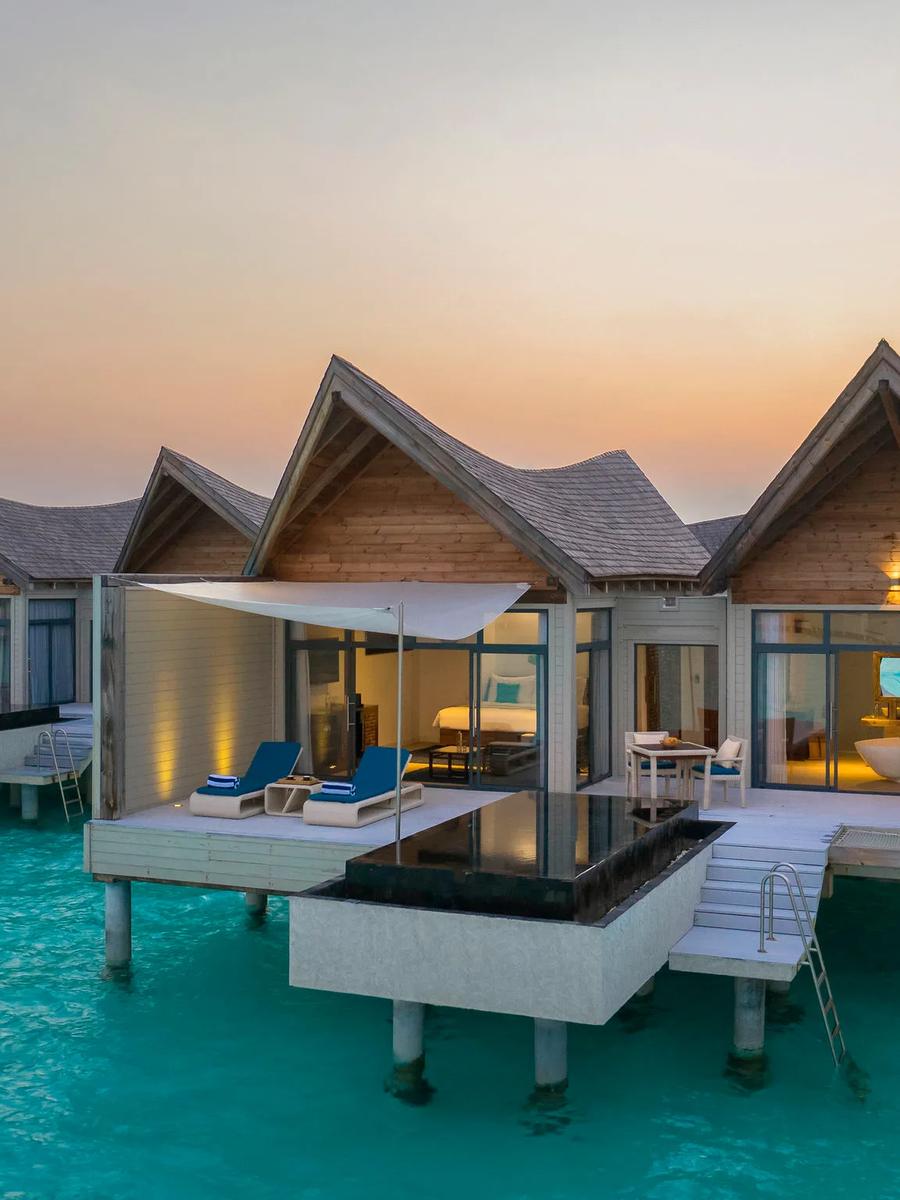 Exterior view of an overwater pool villa at dusk, gabled wood roofs and a private plunge pool on the deck, steps leading down into the lagoon
