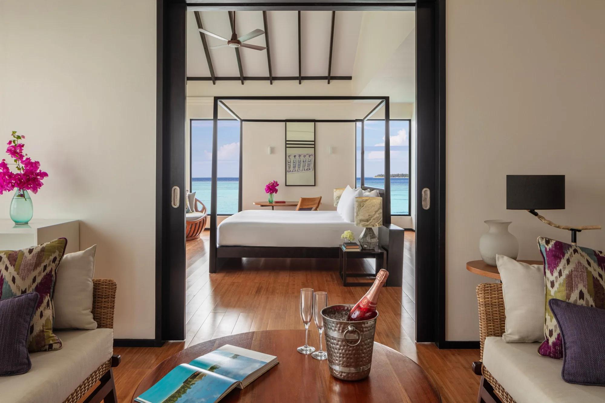A champagne bucket and two flutes on a coffee table in an Over Water Pool Suite's living area, with a four-poster bed and turquoise sea visible through the room beyond
