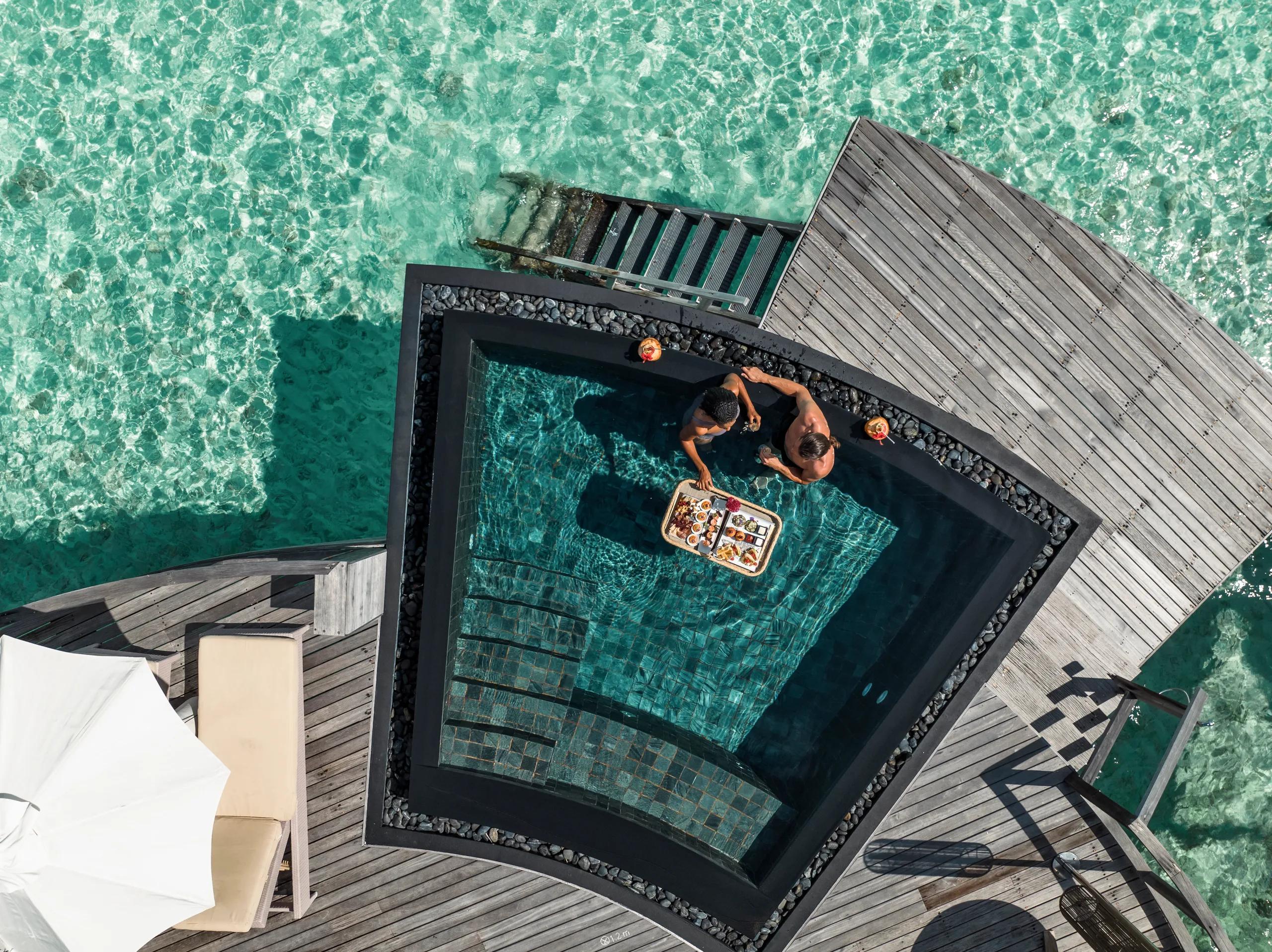 Aerial view directly above a Water Villa's plunge pool, two guests standing in the water with a floating breakfast tray, the turquoise lagoon surrounding the deck