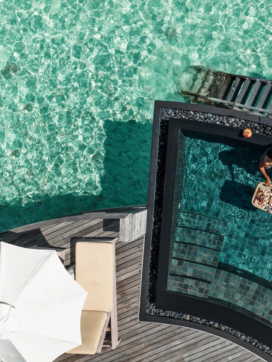 Aerial view directly above a Water Villa's plunge pool, two guests standing in the water with a floating breakfast tray, the turquoise lagoon surrounding the deck