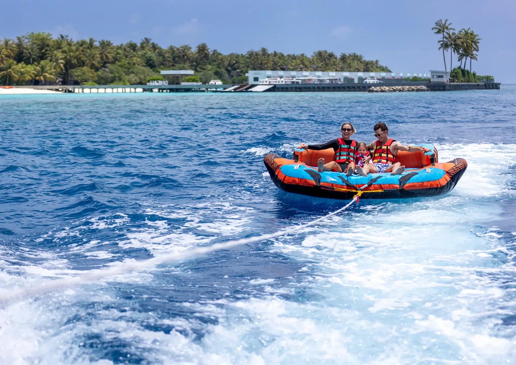 A family of three riding an inflatable towable behind a speedboat, Niva Dhigali's jetty visible behind them
