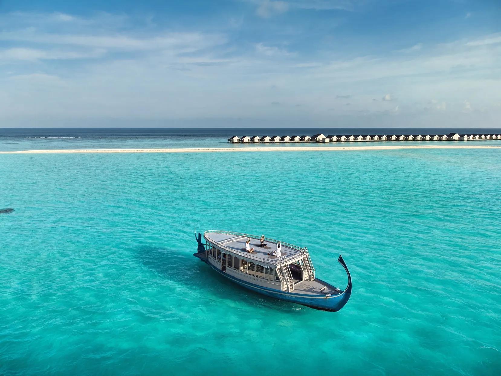 A traditional Maldivian dhoni anchored in the lagoon with guests practising yoga on its rooftop deck, LUX* South Ari Atoll's overwater villa jetty visible in the distance