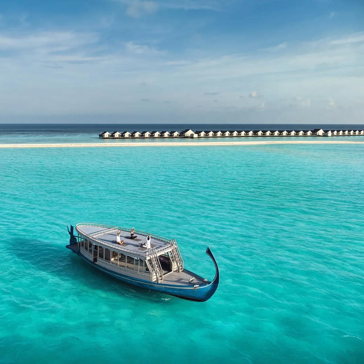 A traditional Maldivian dhoni anchored in the lagoon with guests practising yoga on its rooftop deck, LUX* South Ari Atoll's overwater villa jetty visible in the distance