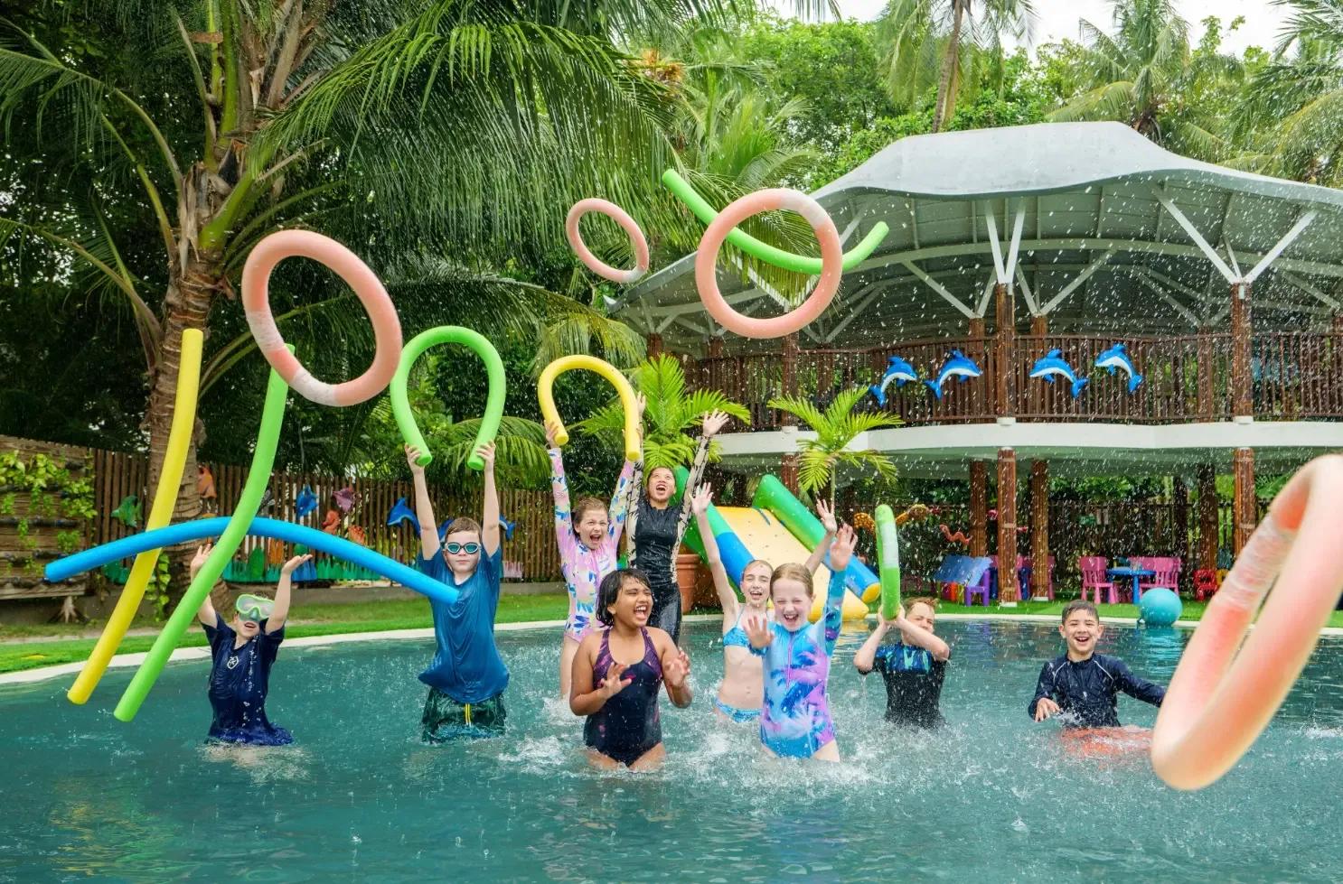 Children playing with pool noodles in Bageecha Kids Club's splash pool, the two-storey club building behind them