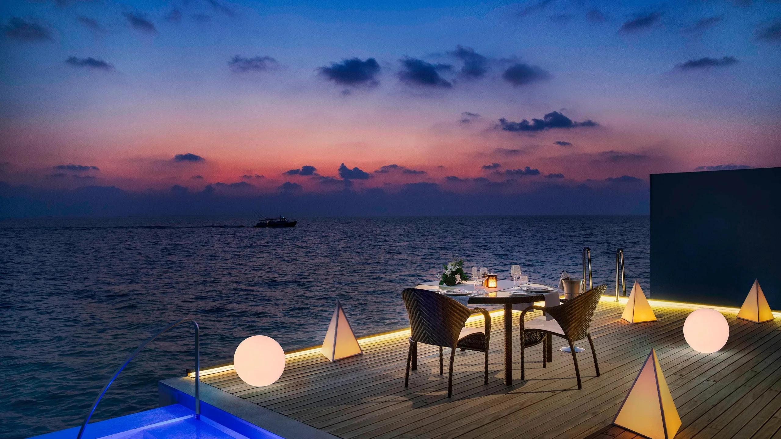 A private dinner table laid on an overwater deck at dusk, lit by lanterns