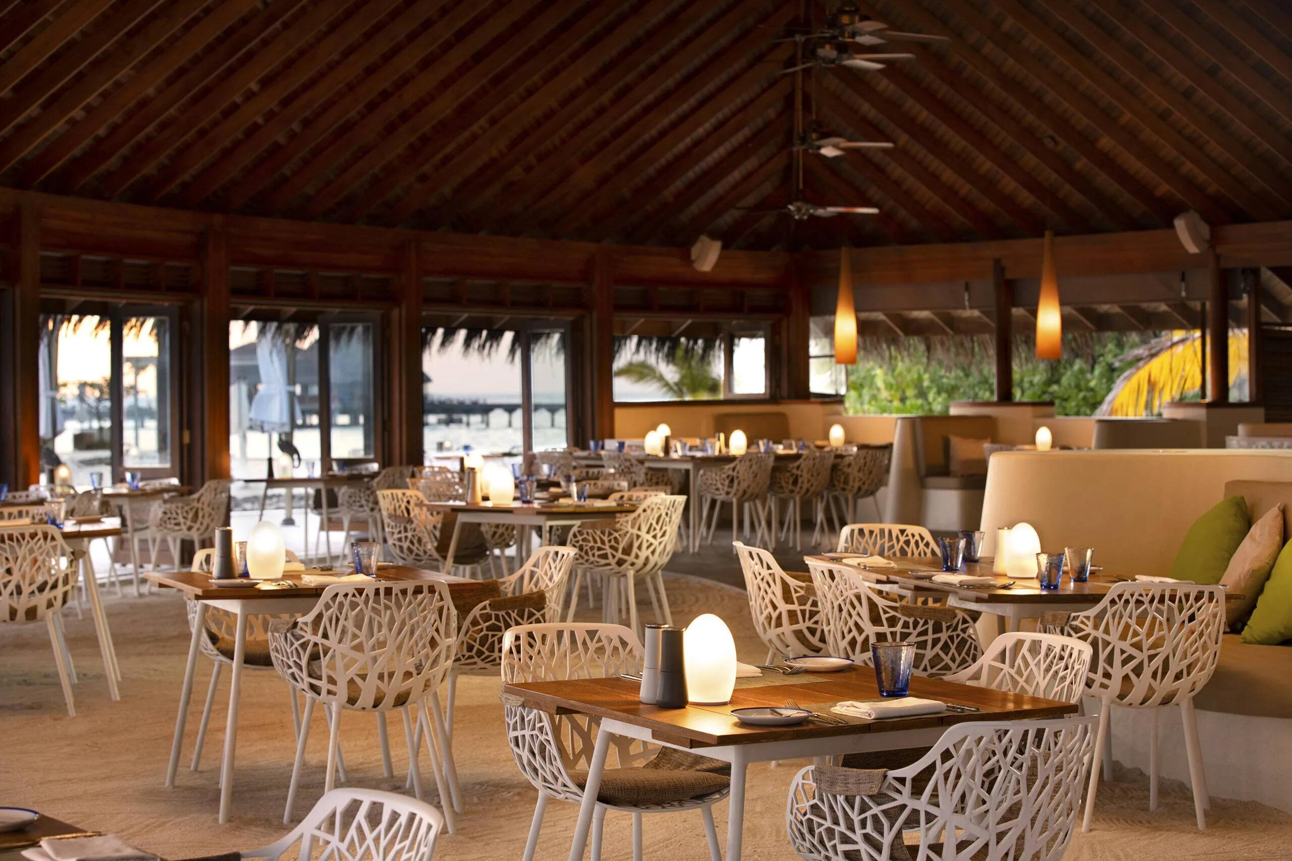 Celsius's dining room at dusk, tables set beneath a high thatched ceiling with the jetty visible through open doors