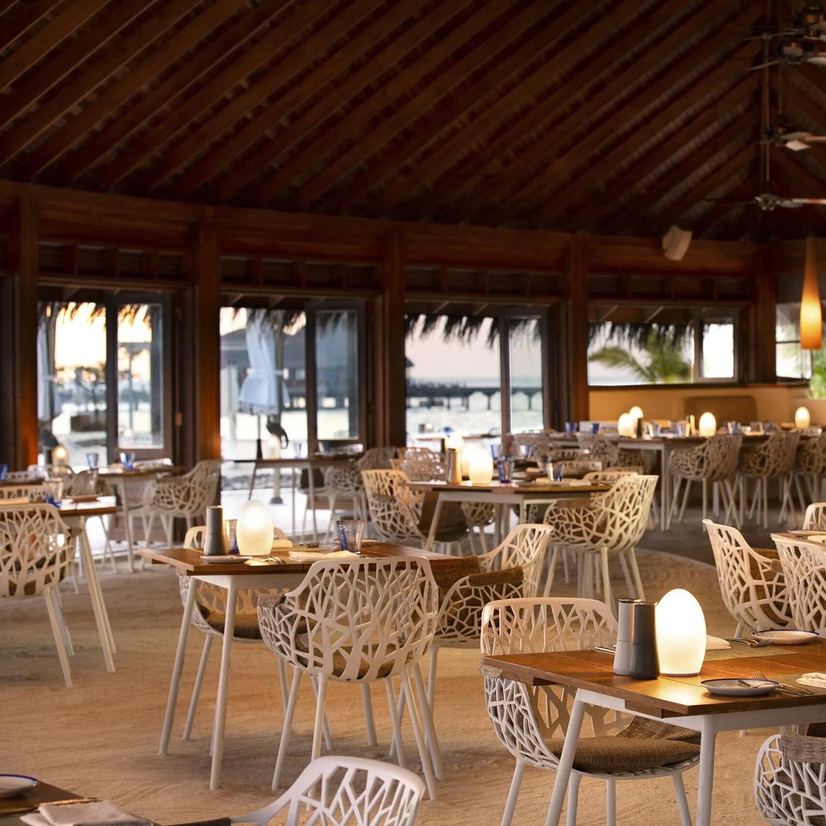 Celsius's dining room at dusk, tables set beneath a high thatched ceiling with the jetty visible through open doors