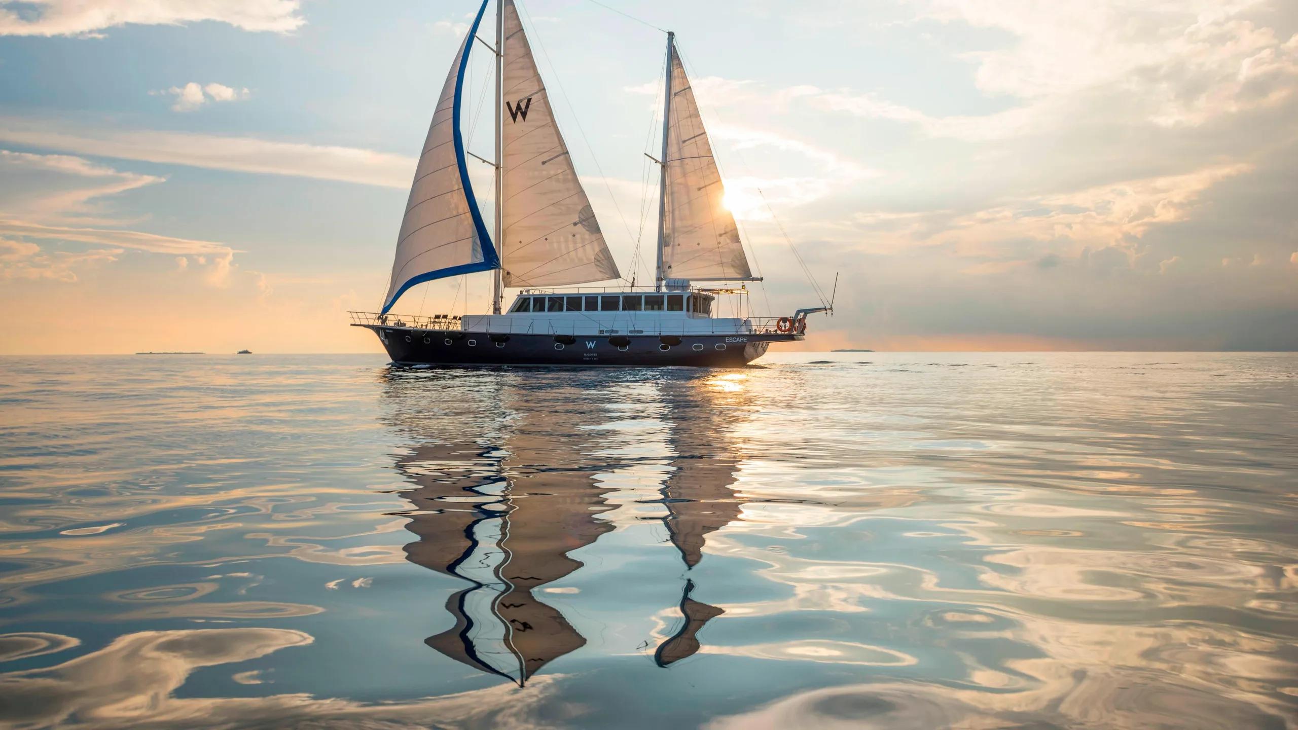 W Maldives' own sailing yacht under full sail at sunset, reflected in the still water