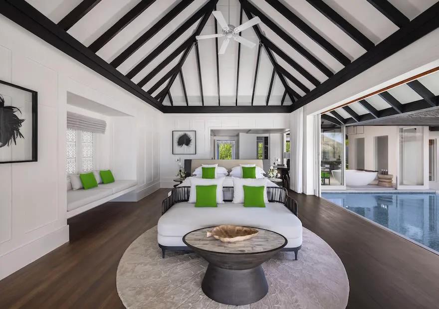A bedroom with a vaulted thatch ceiling, green accent cushions, and glass doors opening onto a private pool deck