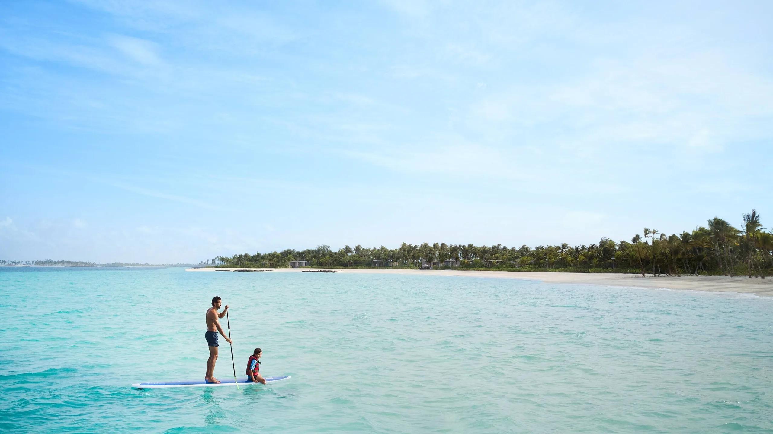 A father and child paddleboarding across shallow lagoon water toward a palm-lined island