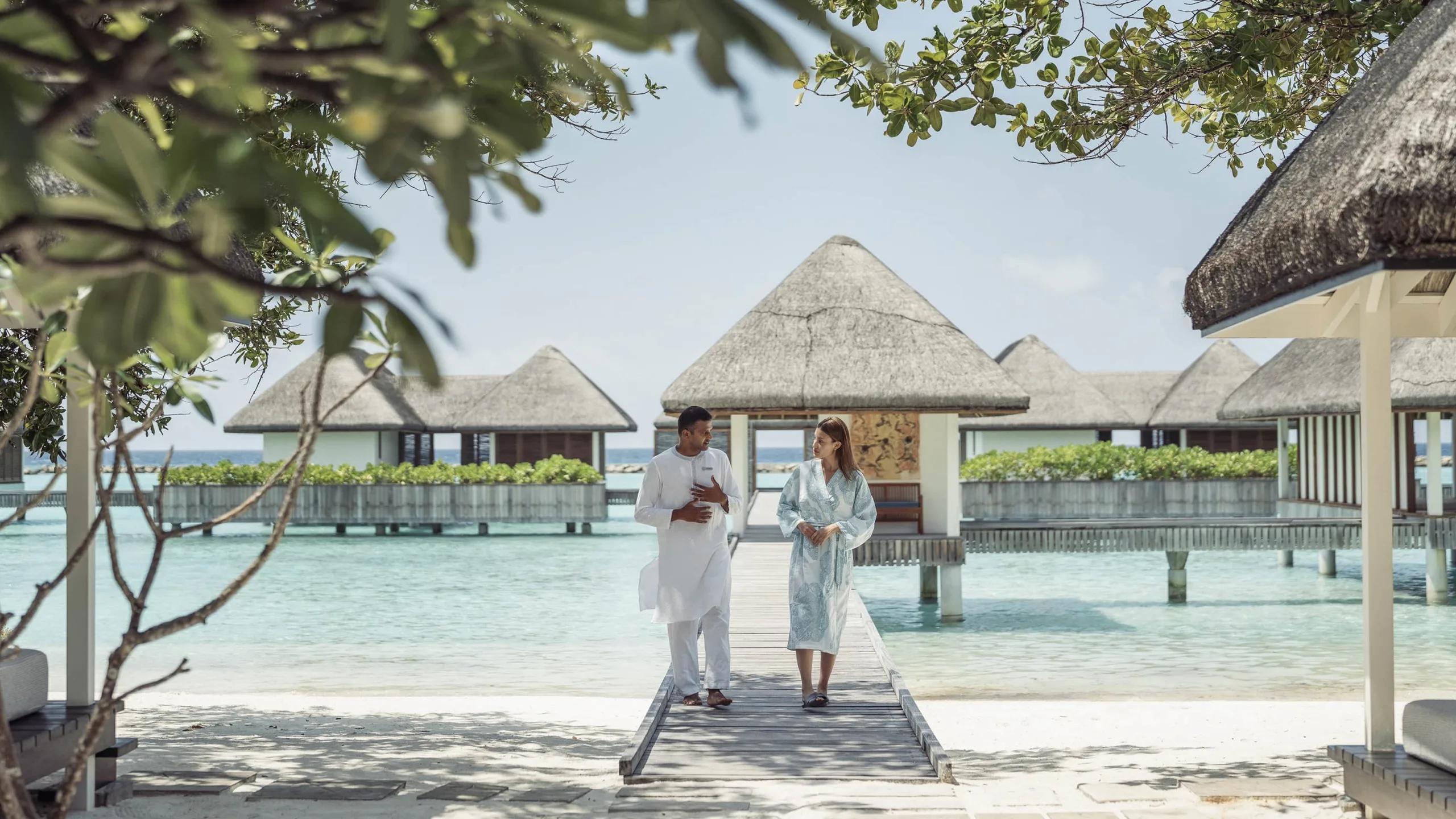 A spa host in white escorting a guest in a robe along the overwater boardwalk toward AyurMa's pavilions